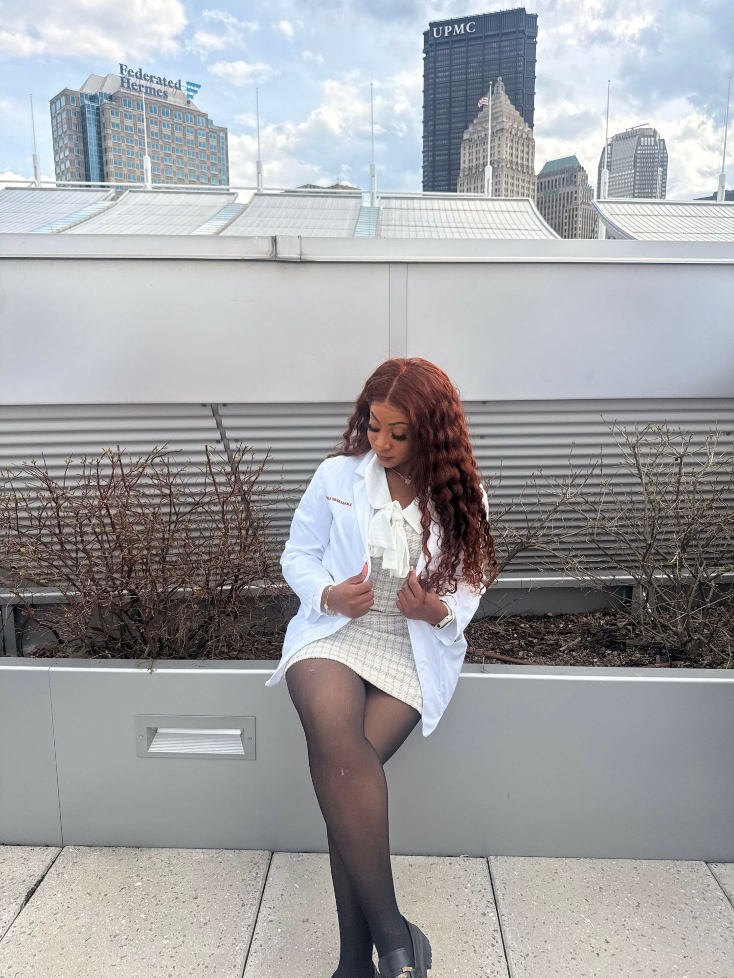 Woman with long hair in white outfit and black tights standing outdoors on a city rooftop.