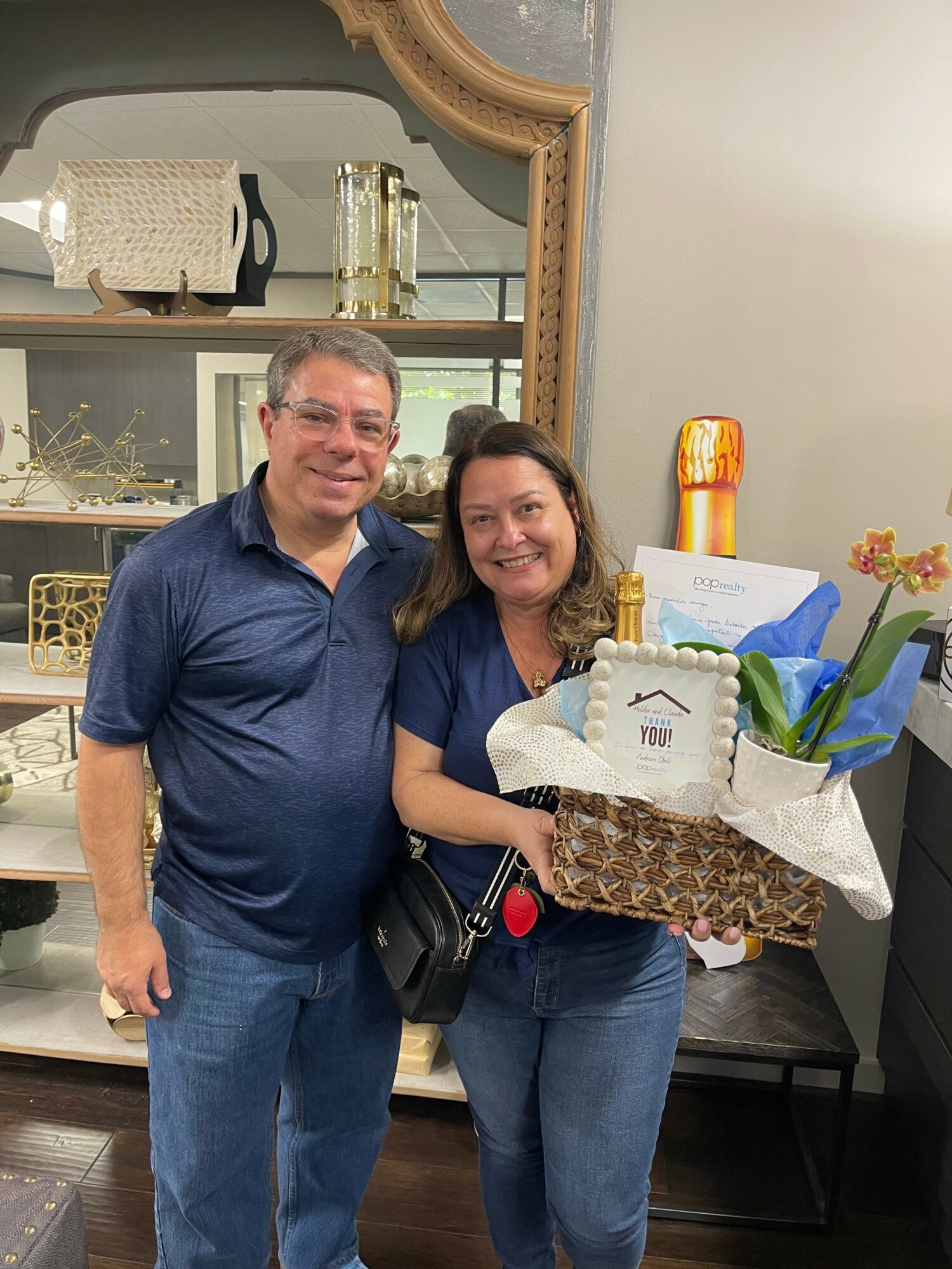 Two people, a man and a woman, stand indoors holding a gift basket with flowers and a card, smiling at the camera.