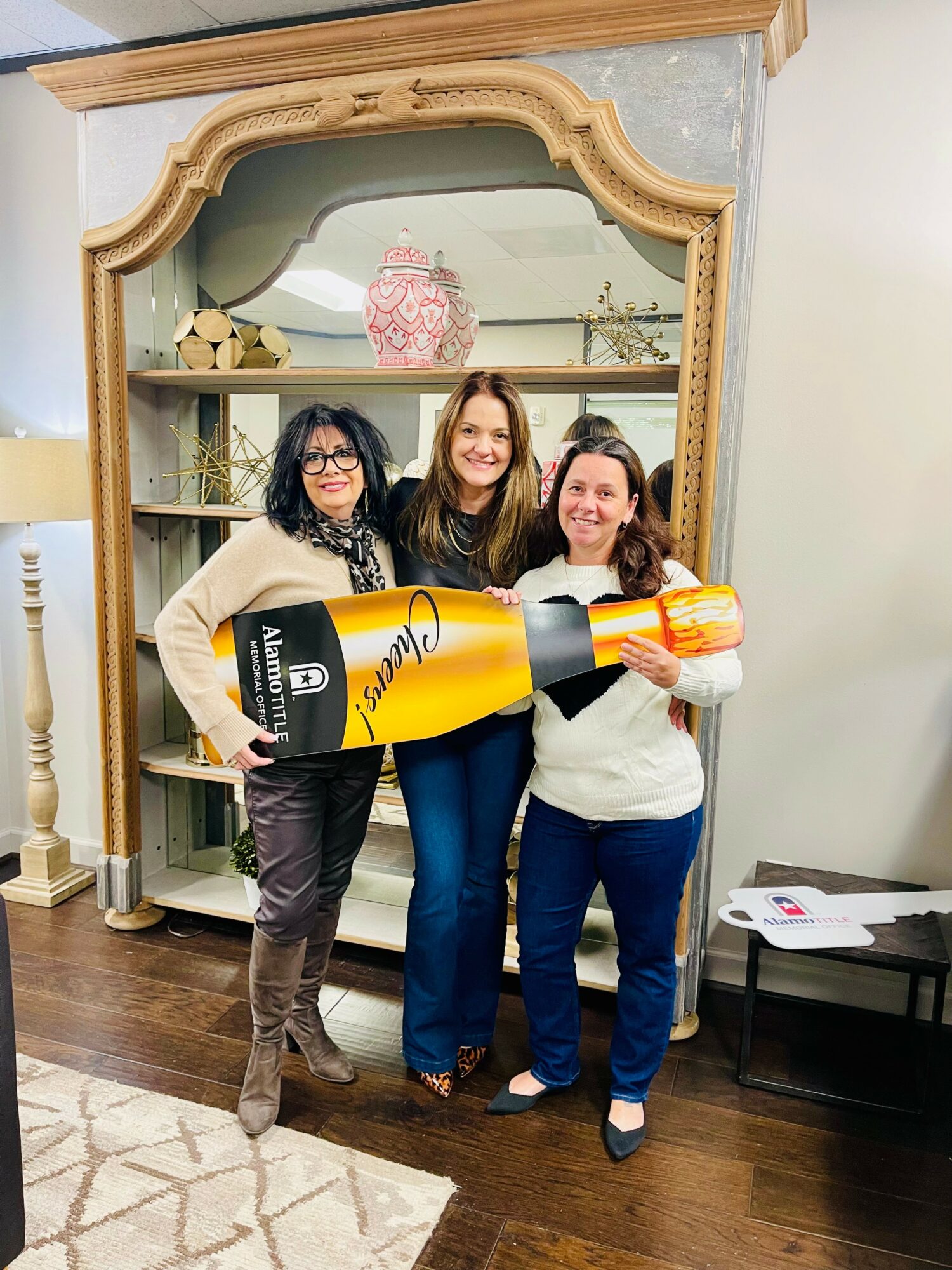 Three women stand together holding a large yellow and black rocket-shaped object in front of a mirror with a wooden frame, smiling.