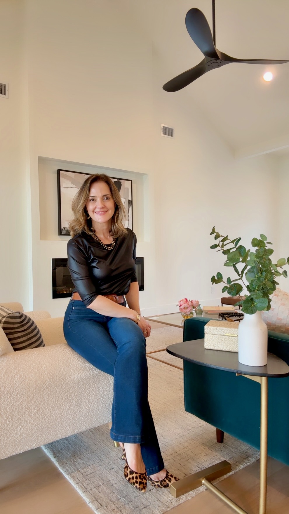 Woman sitting on a sofa in a living room with a plant and decorative items, smiling at the camera.