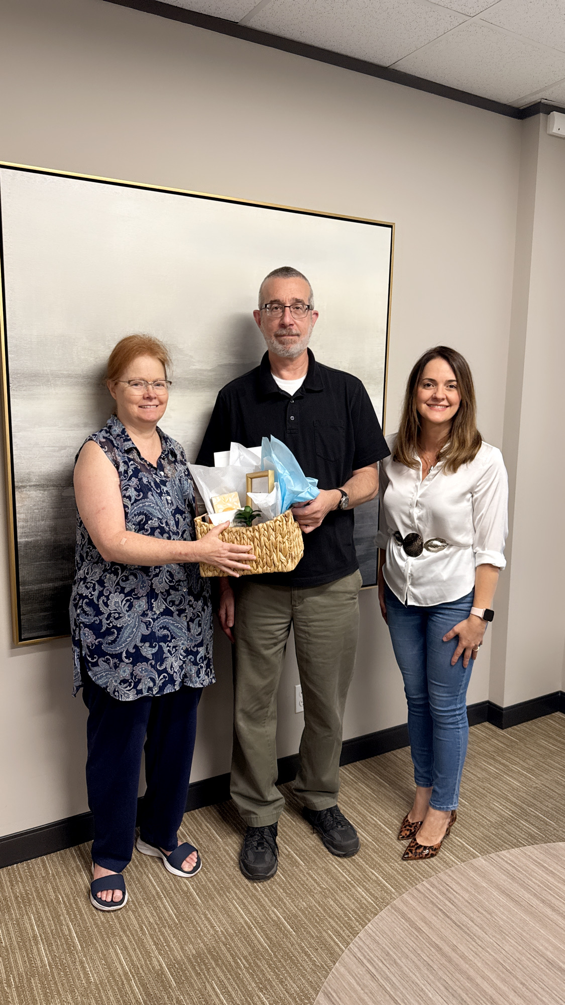 Three people standing indoors in front of a whiteboard, holding a gift basket, smiling.