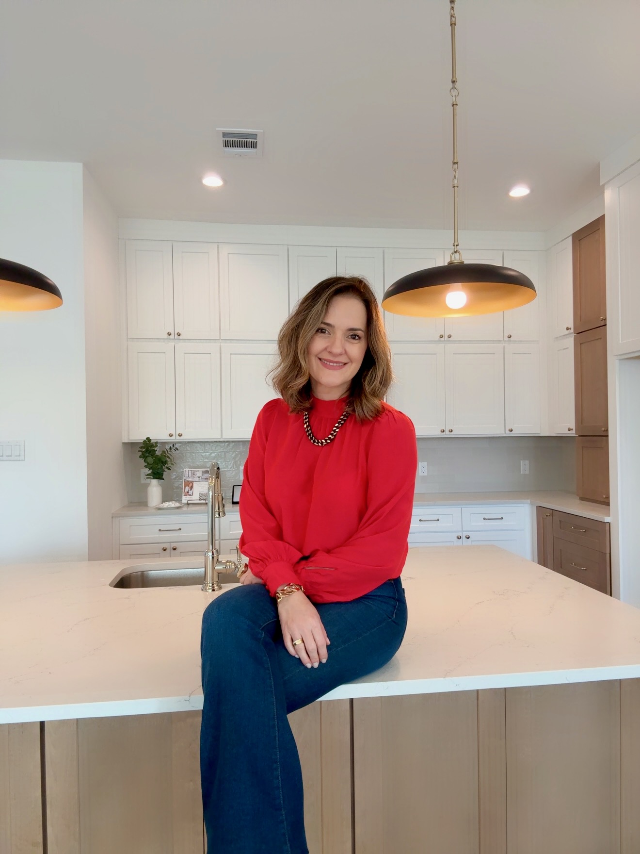 Woman with shoulder-length hair in a red sweater and blue jeans sitting on a kitchen island, smiling.