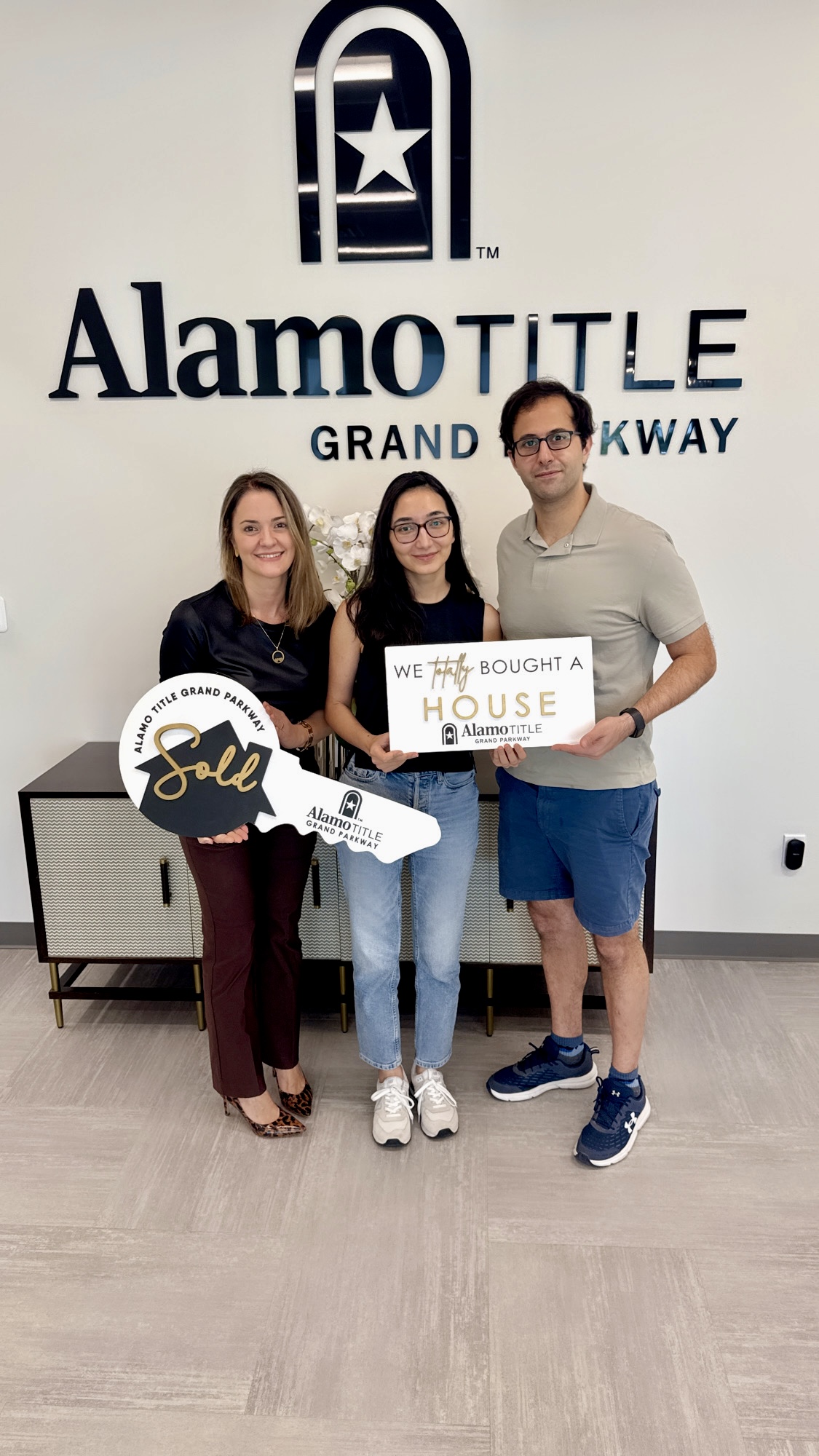 Three people standing indoors holding signs in front of a wall with 'Alamo TILE' logo and text.