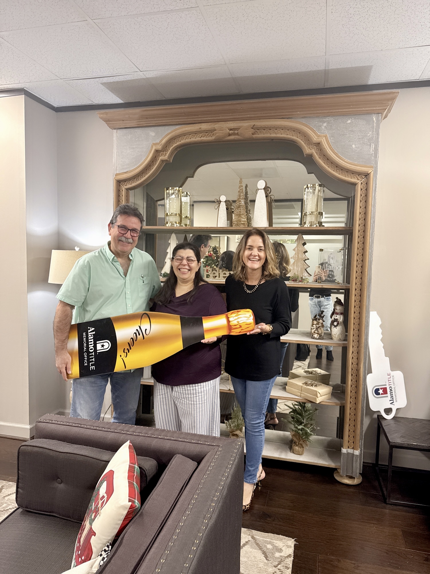 Three people standing indoors holding a large yellow and black object, smiling, with a decorative mirror and shelves behind them.