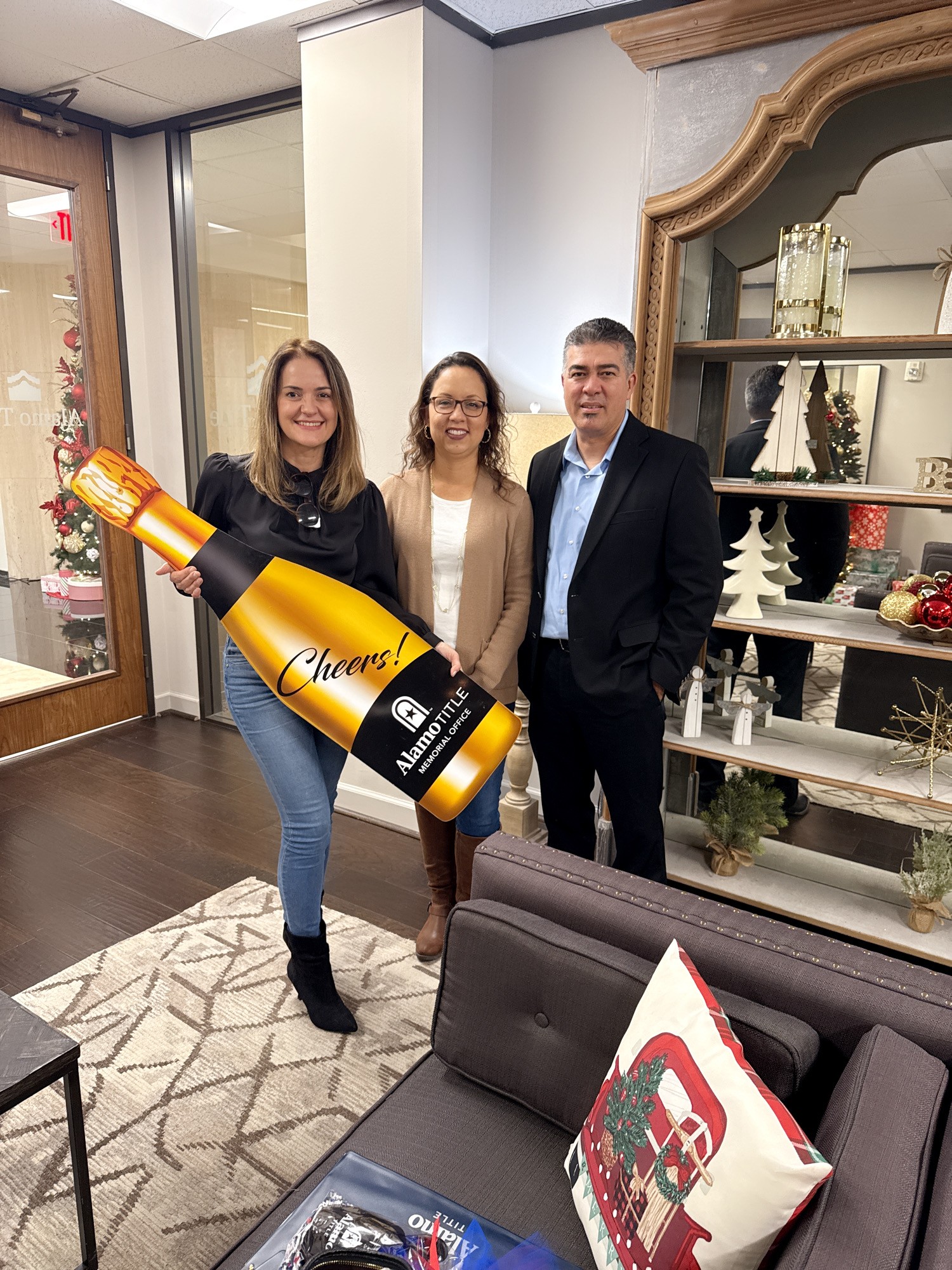 Three people standing indoors, smiling, with a large champagne bottle decoration, in a room with shelves and holiday decorations.