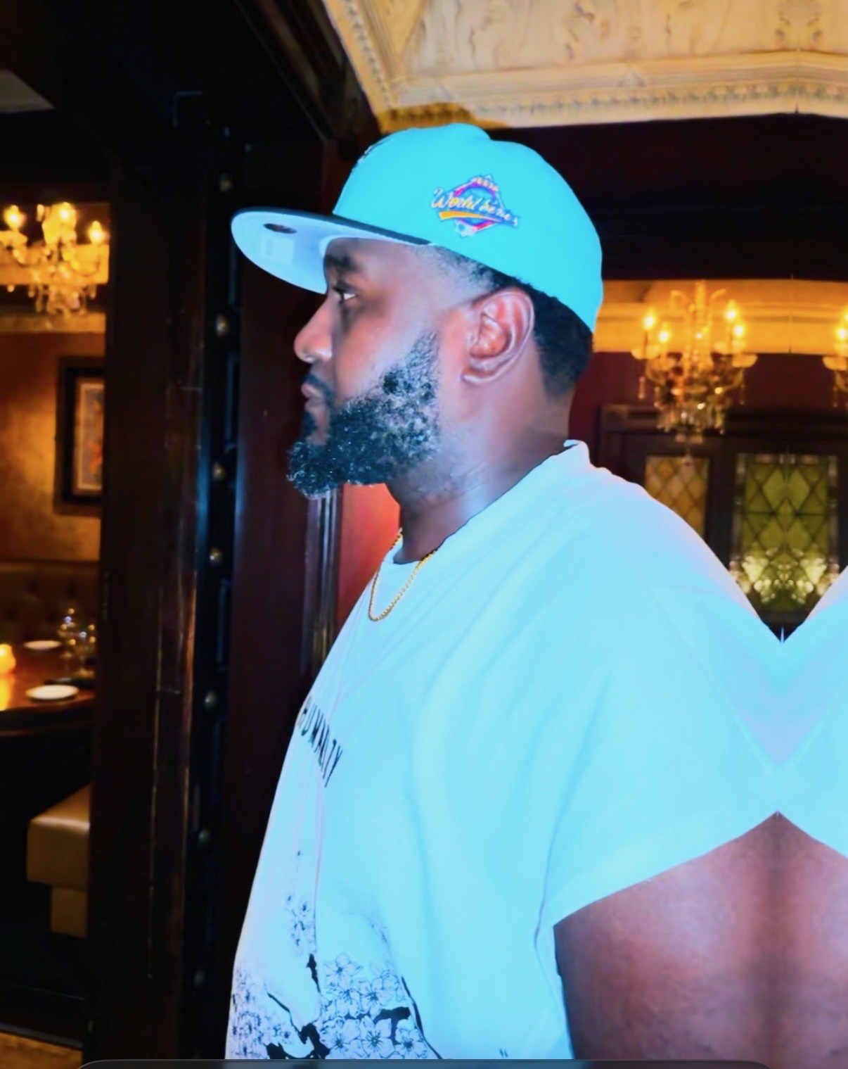 Side profile of a man wearing a blue cap and white T-shirt, indoors with warm lighting and chandeliers.
