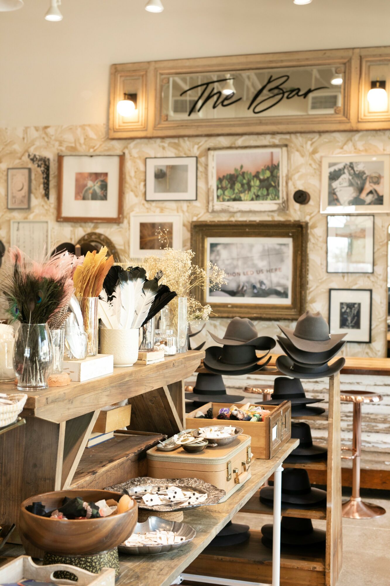 Interior of a cafe with a wall decorated with framed pictures and a sign reading 'The Bar'. Wooden shelves hold hats and decorative items.