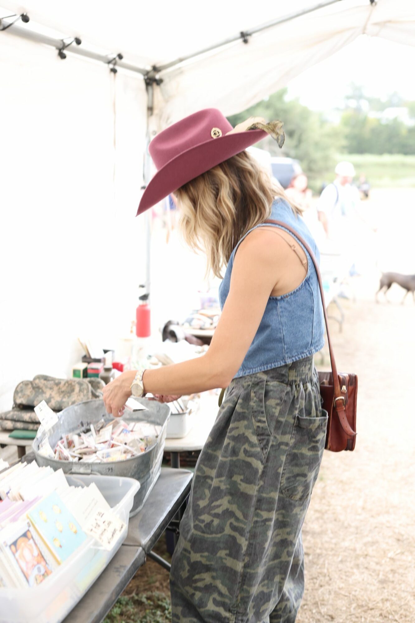 Woman wearing a pink hat and denim vest at outdoor market stall, reaching for items on table, tent in background.