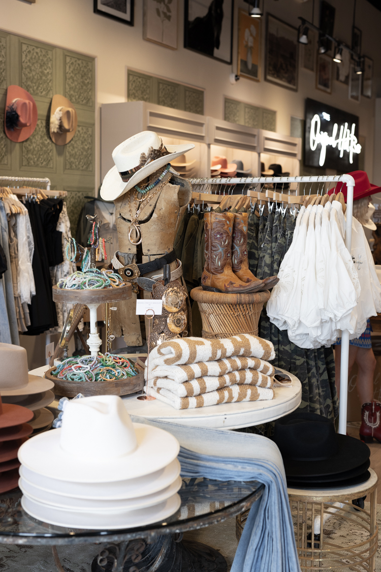 Display of hats, clothing, and accessories in a boutique store with shelves and racks, and a neon sign in the background.