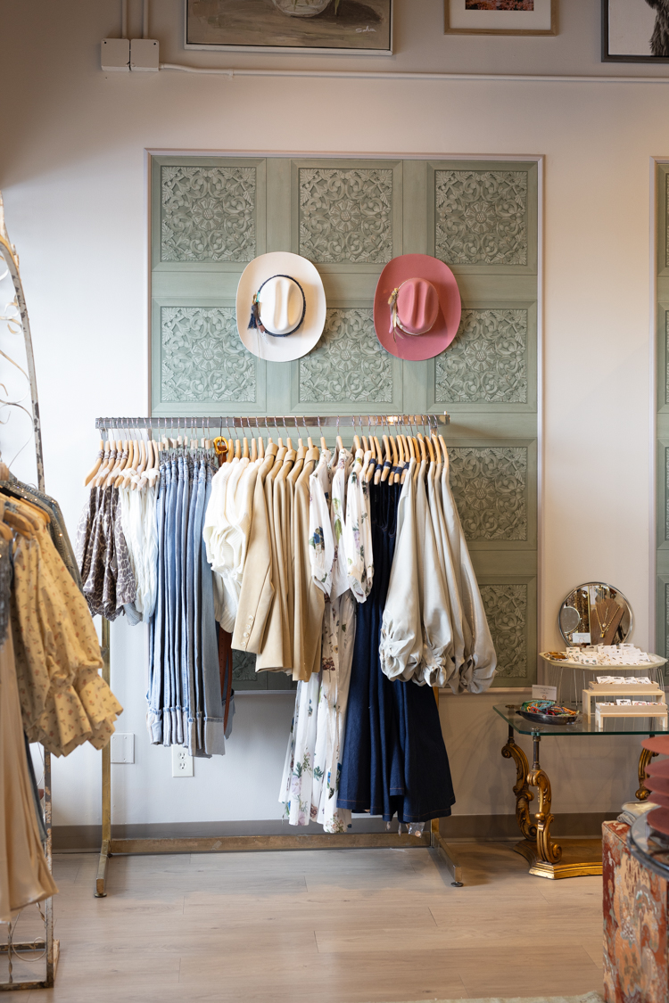 Clothing rack with white and dark garments, hats on a decorative wall, and a side table with a clock and books.