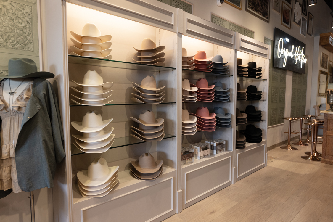 Display of hats and caps on shelves in a store, with clothing on the left and a neon sign on the right, wooden floor.