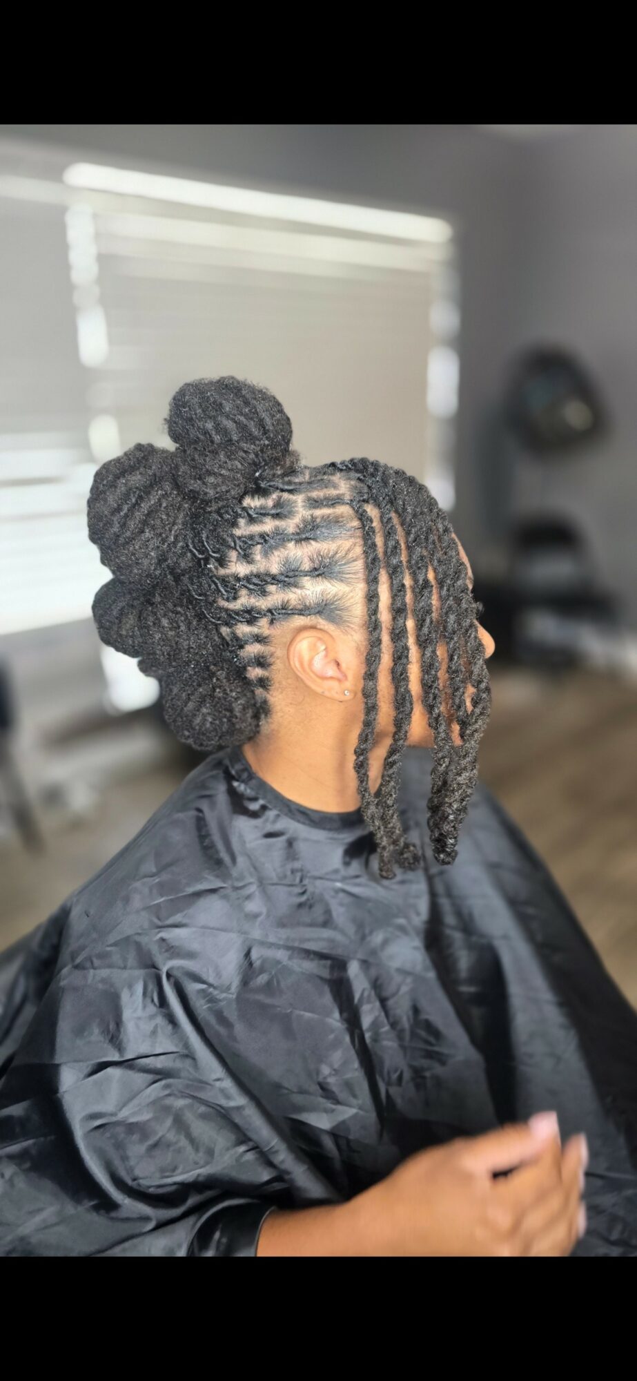 Person with braided hairstyle and large bun, sitting in a salon chair, wearing a black cape.