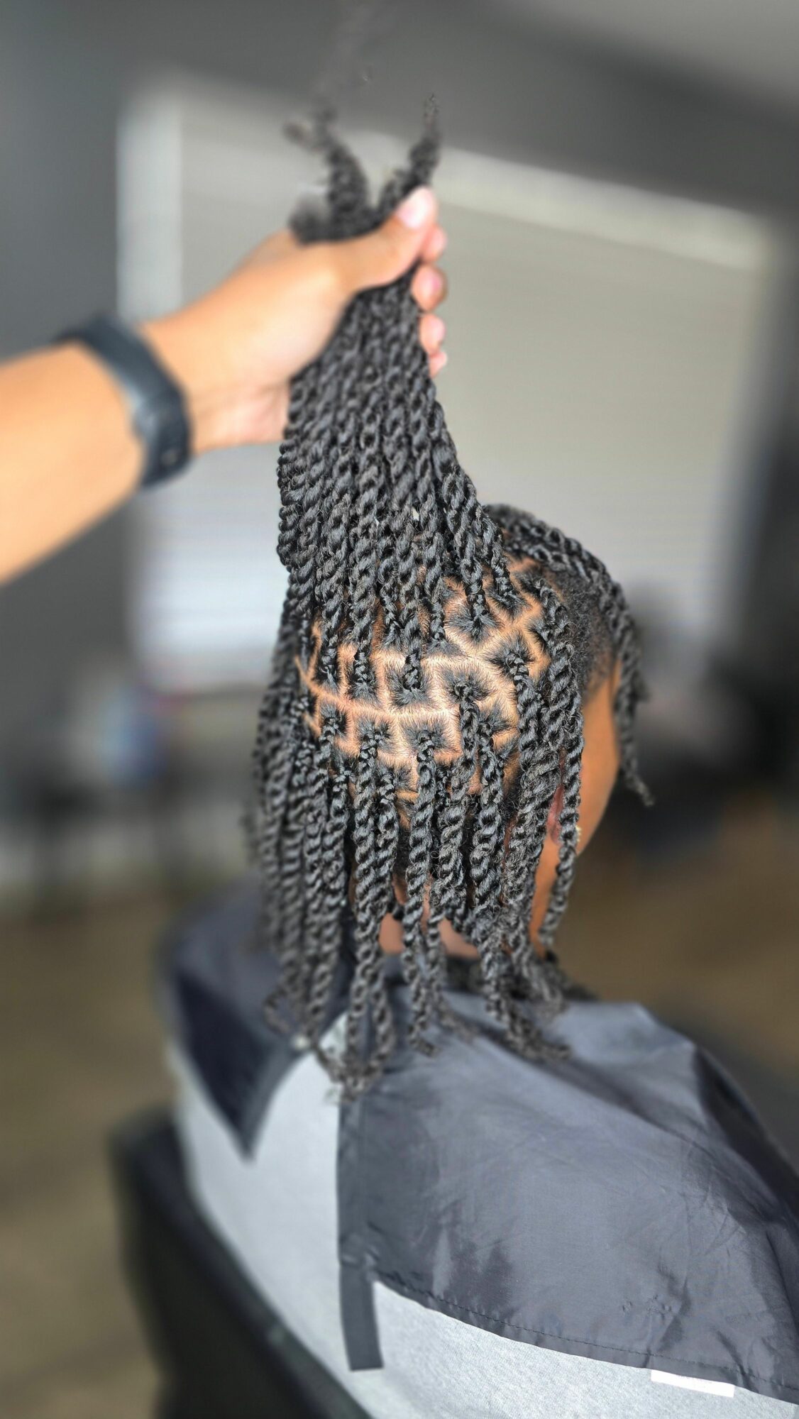 Person with braided hair, wearing a gray and black jacket, holding a section of hair.