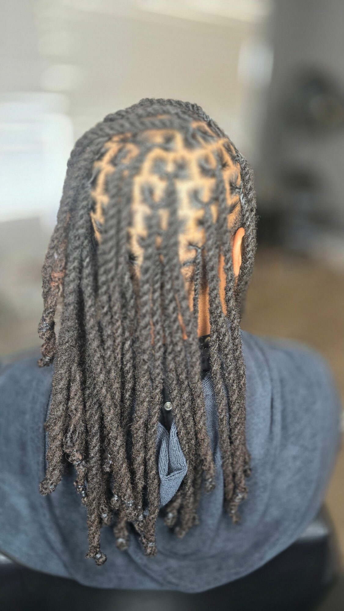 Person with long dreadlocks, some with intricate patterns, wearing a blue shirt, viewed from behind.