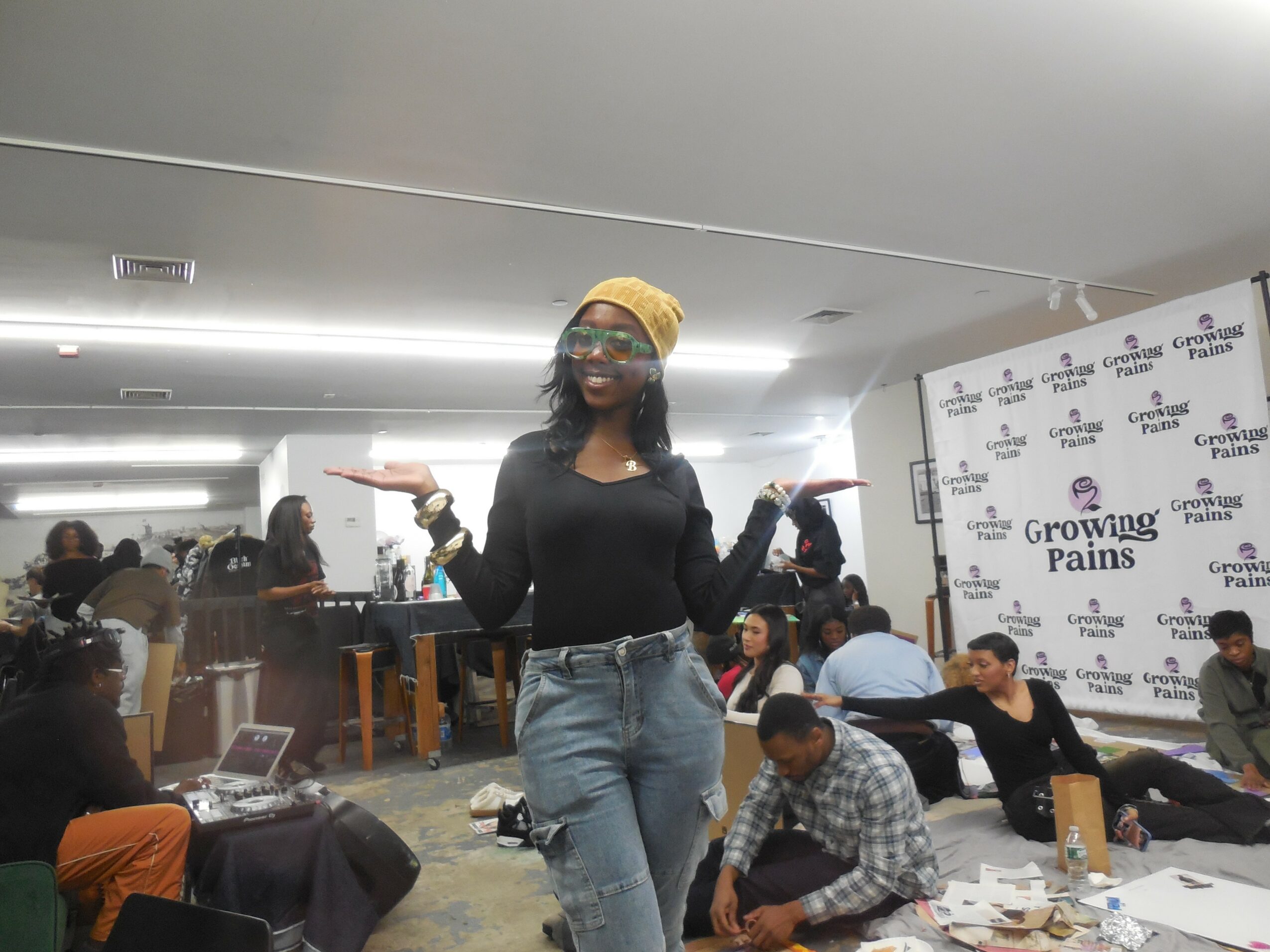 Woman wearing a yellow headscarf and glasses, standing with arms outstretched in a room with people working at tables.