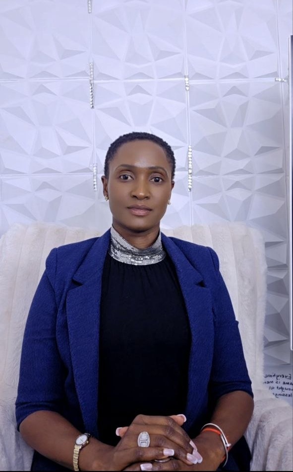 Woman with short hair wearing a blue blazer and black top, sitting in front of a textured white wall.