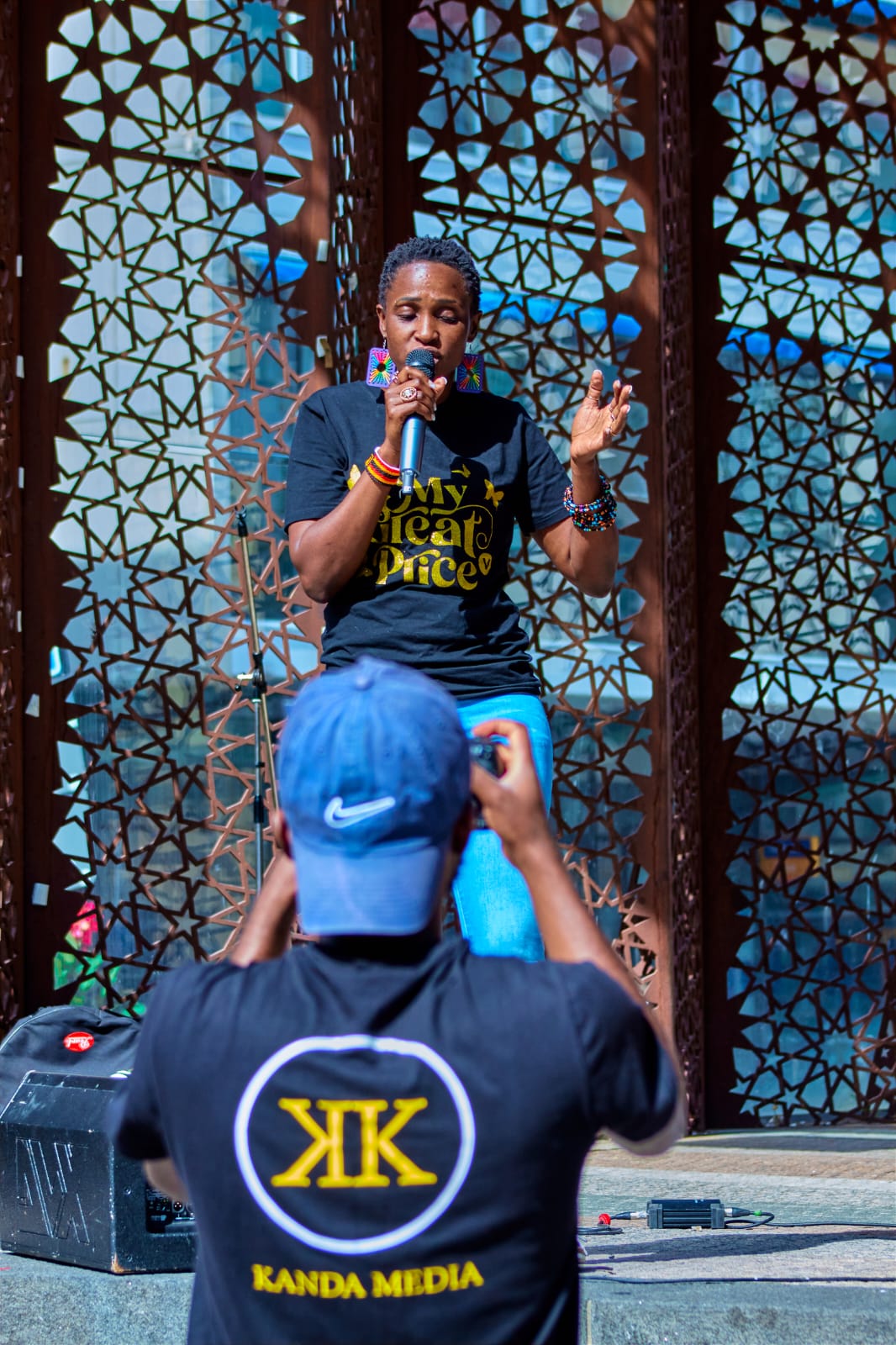 Person speaking into microphone on stage with decorative background, audience member in foreground wearing a blue cap, Kanda Media logo visible.