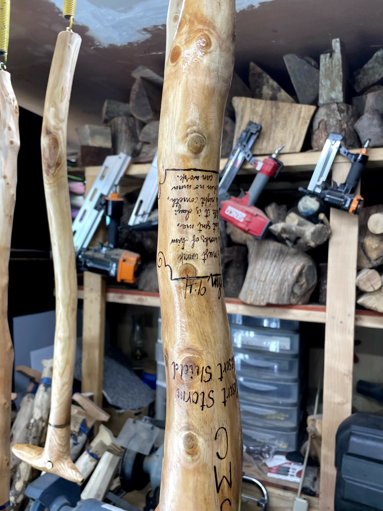 Wooden stick with handwritten text, surrounded by other sticks and woodworking tools in a workshop.