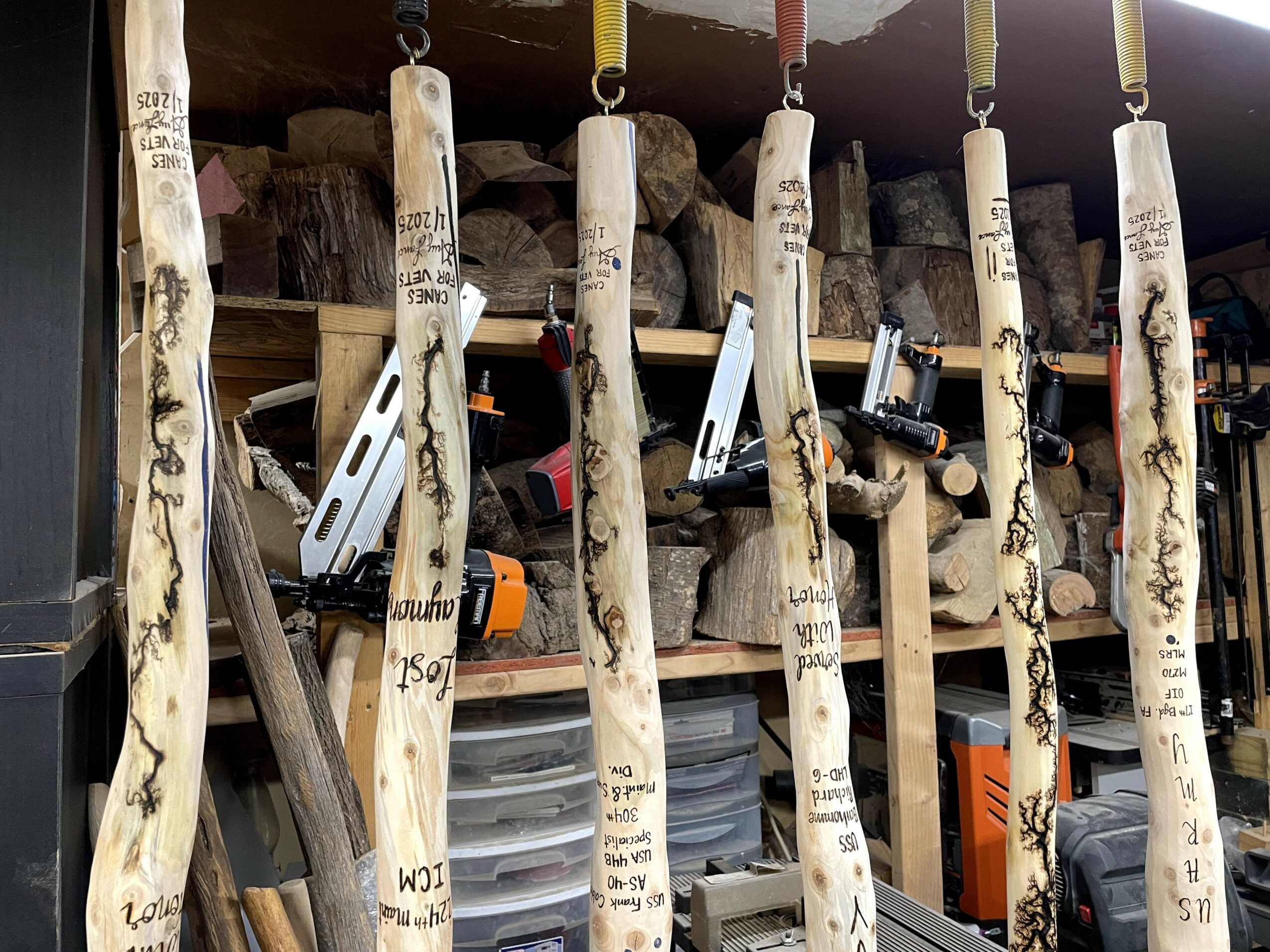 Several wooden sticks with handwritten notes hang from hooks in a workshop, with tools and wood in the background.