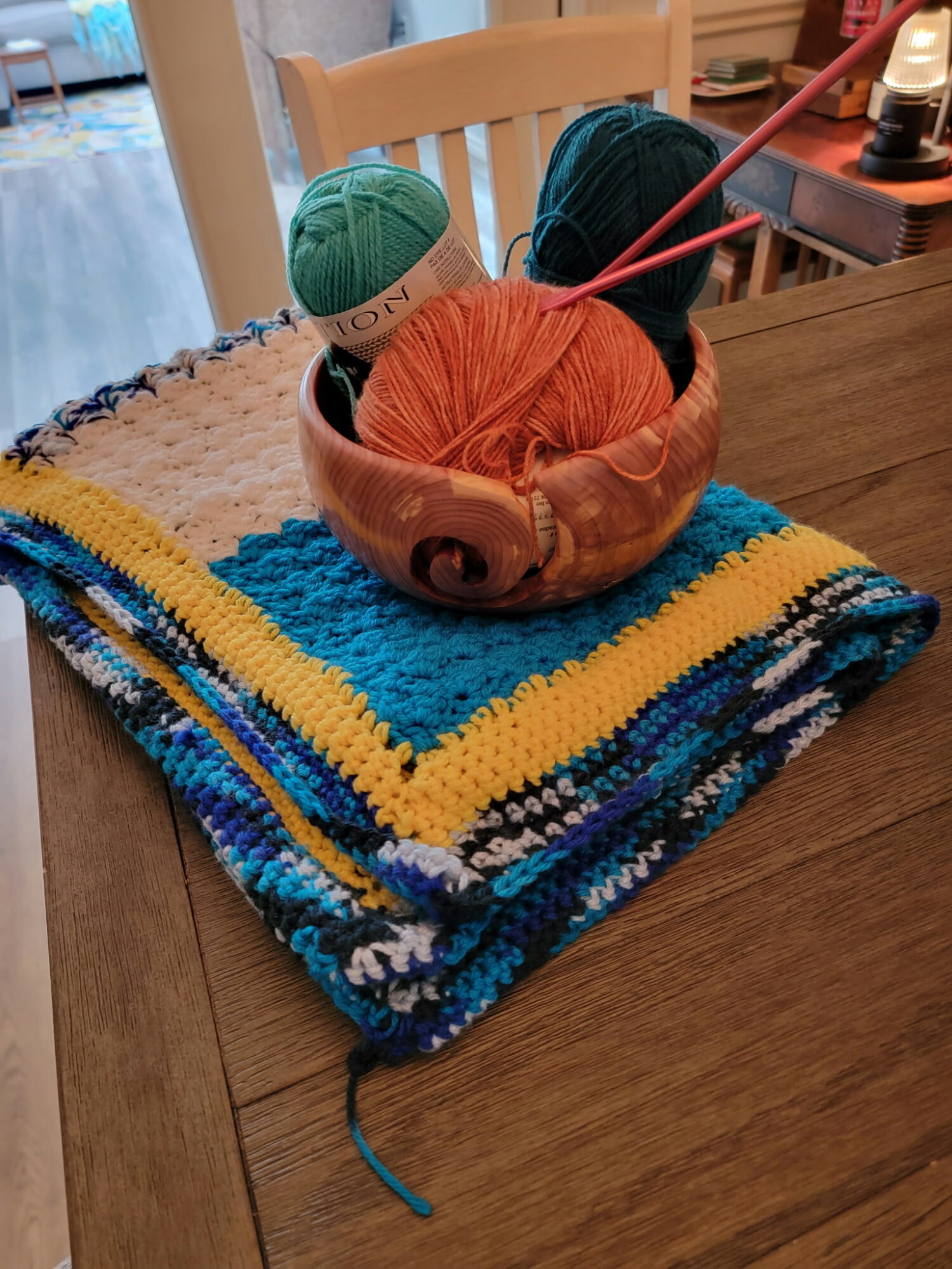 A bowl with yarn balls and knitting needles on a table, placed on a colorful knitted blanket.