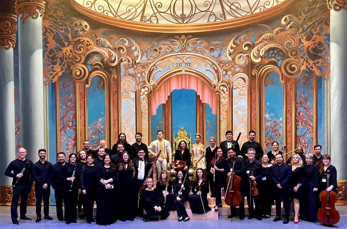 Group of performers on stage with ornate, colorful backdrop and a golden throne, some holding musical instruments, in a theater setting.
