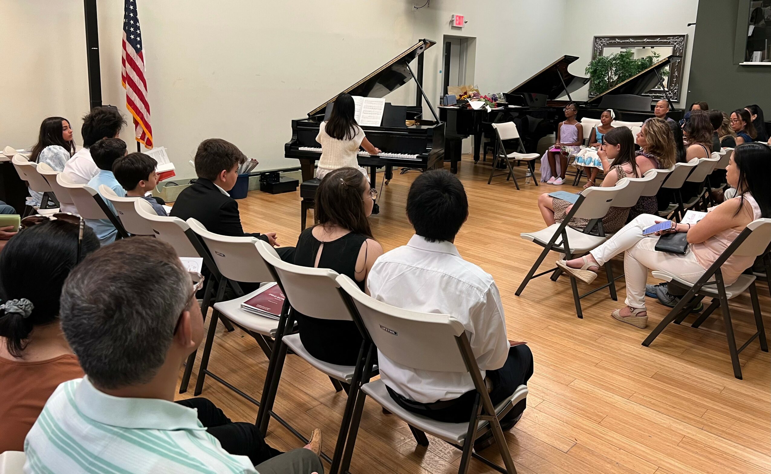 Children and adults seated in chairs facing a stage with two pianos, an American flag, and a plain wall background.