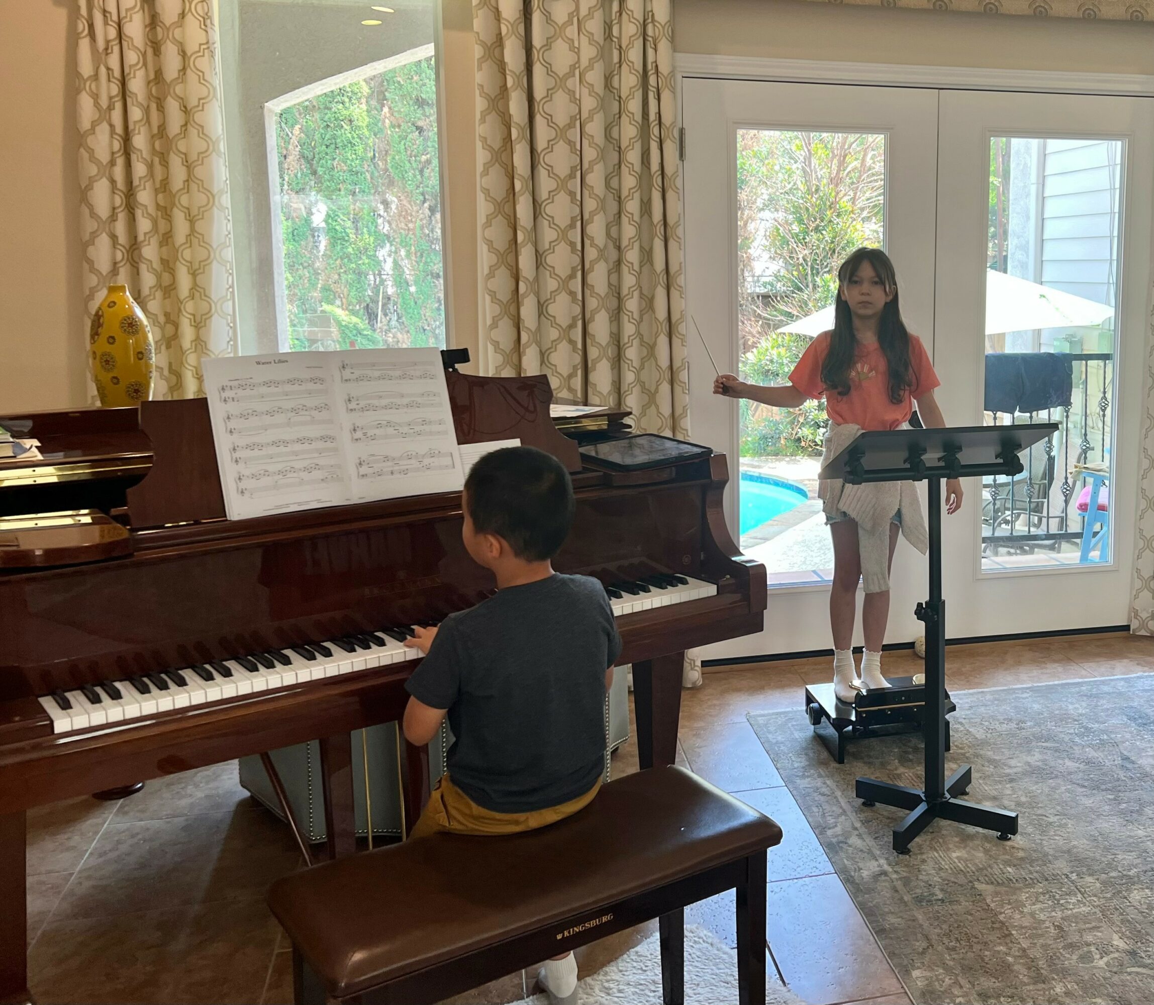 A boy plays piano while a girl sings into a microphone in a sunlit room with large windows and curtains.