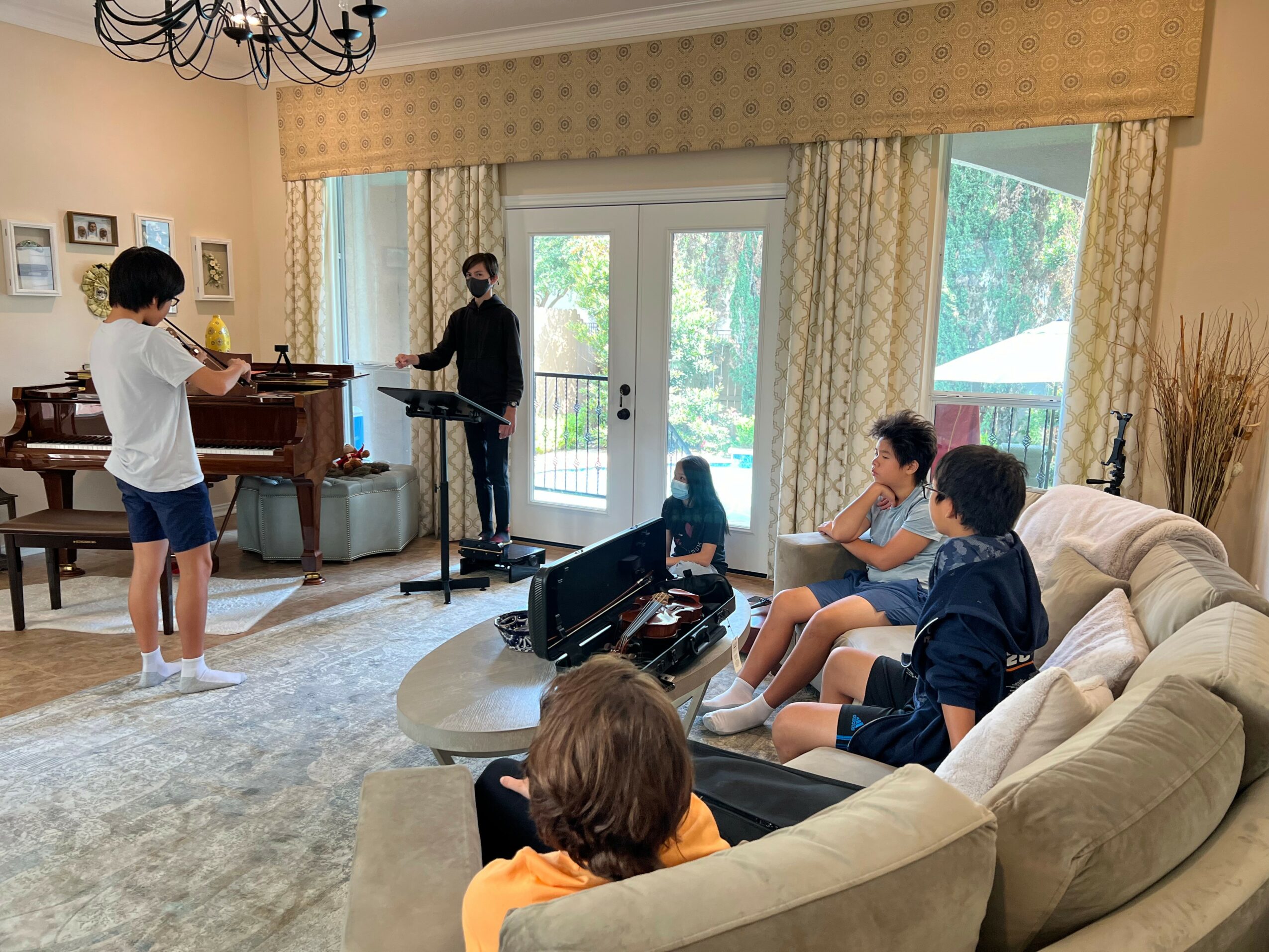 Group of five people in a living room, some sitting on a sofa, others standing near a piano, with large windows and curtains.