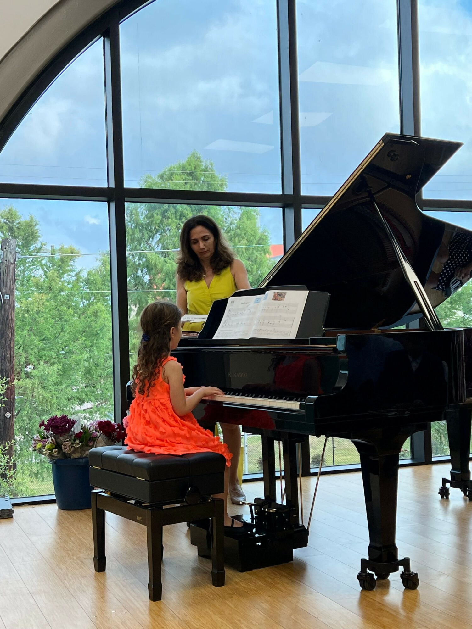 Two women sit at a grand piano in a room with large windows and green trees outside, one in a yellow top, the other in an orange dress.