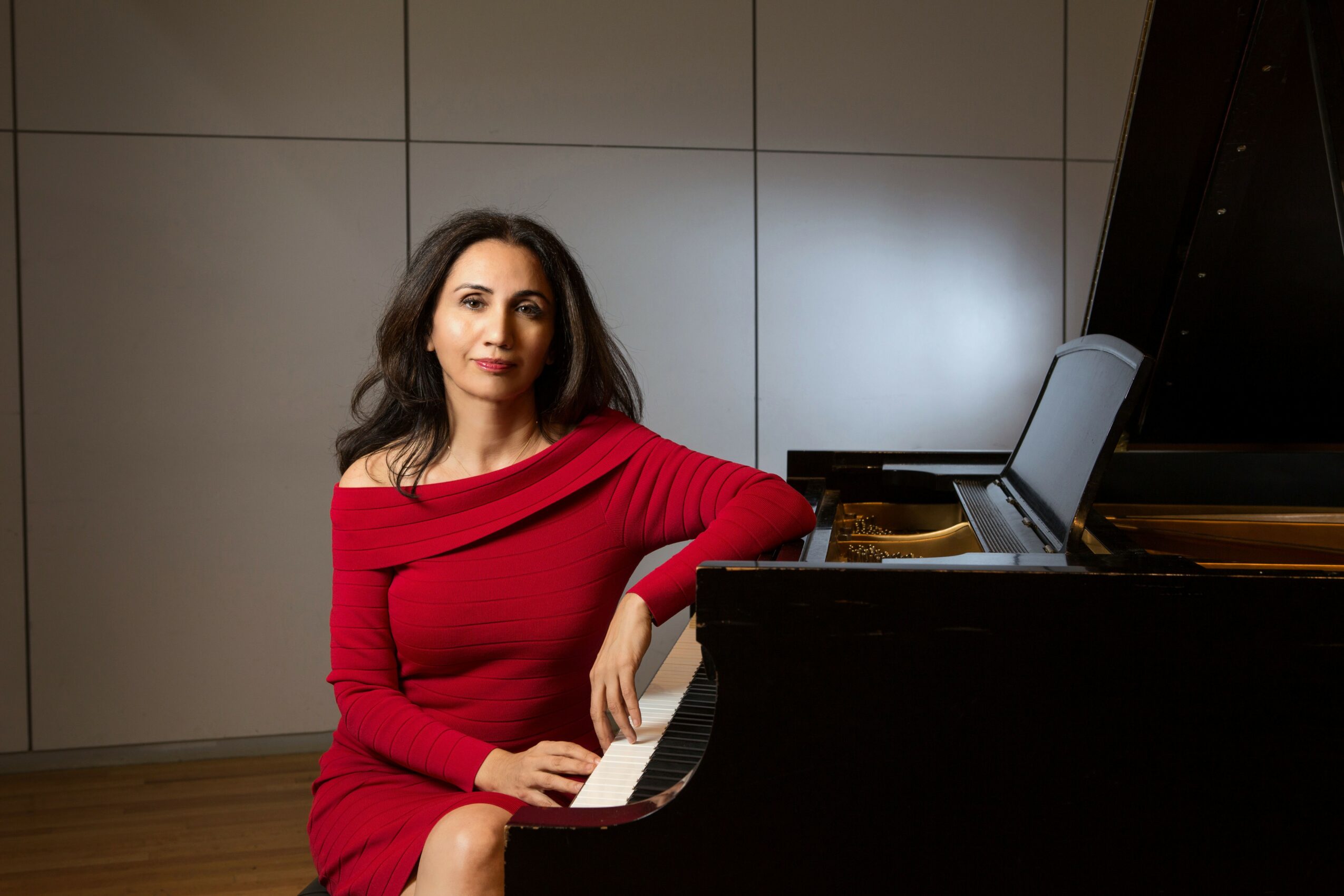 Woman in red dress sitting at a piano with a laptop on top, in a room with plain wall panels.