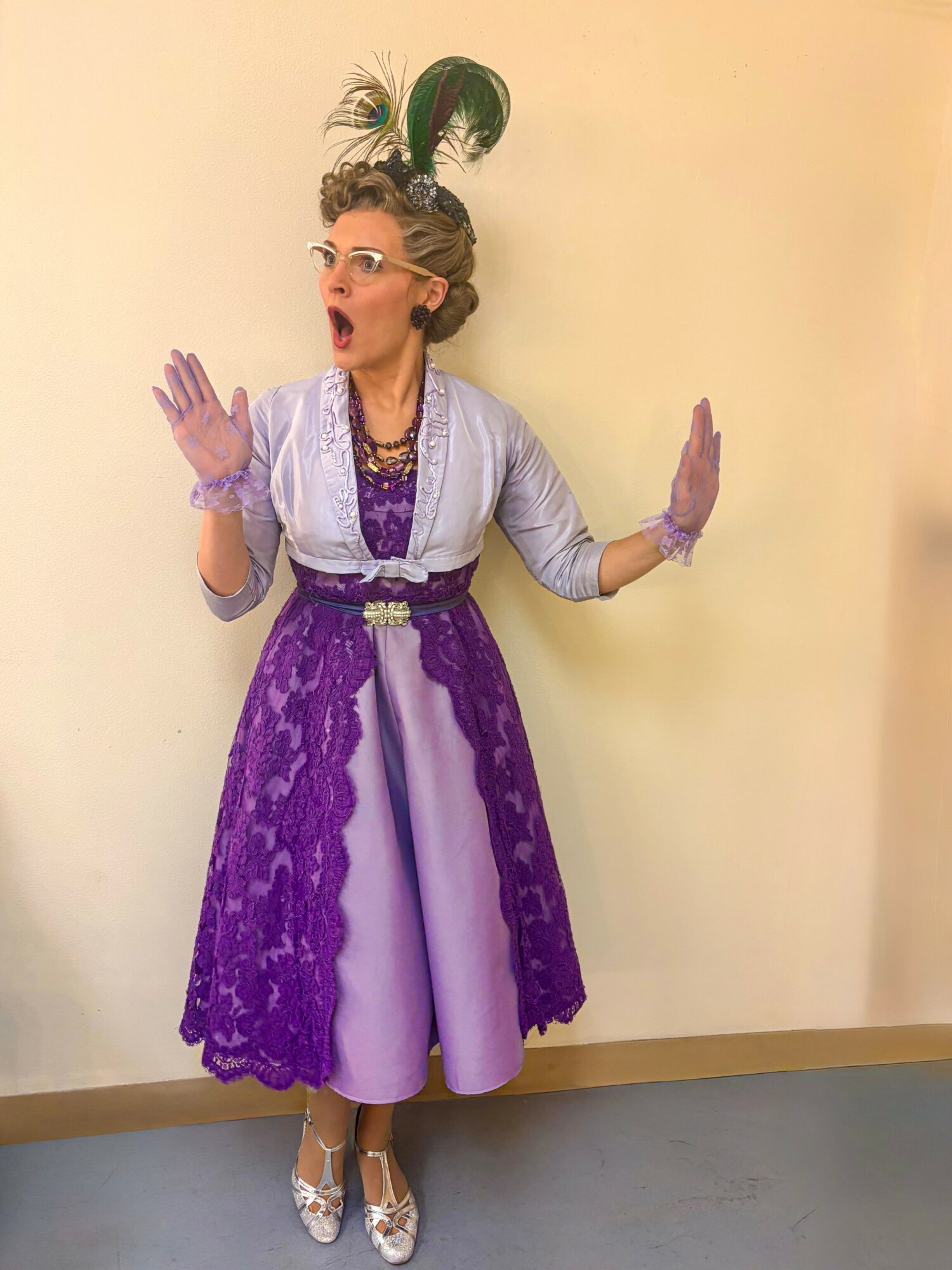 Woman in vintage dress with purple lace overlay, wearing gloves and a feathered headpiece, standing against a plain wall.