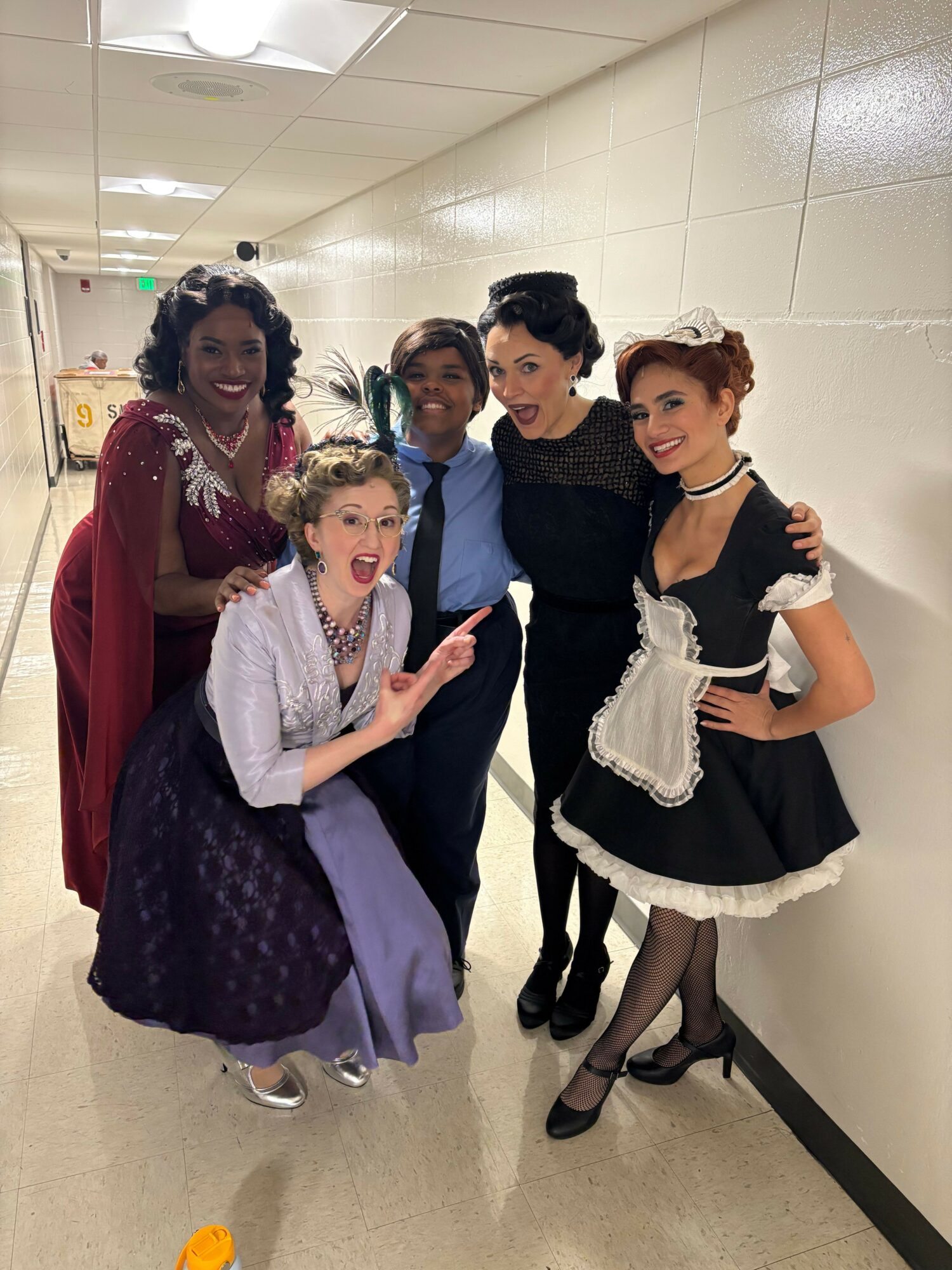 Group of five people in costumes posing in a hallway, some with makeup and accessories, smiling and making gestures.