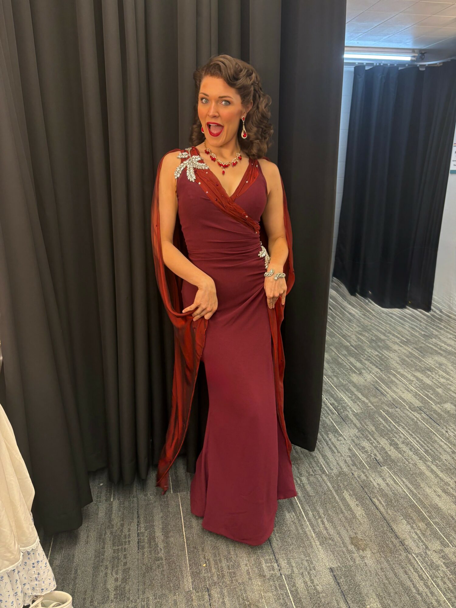 Woman in a burgundy gown with a cape, standing in front of dark curtains, smiling, with a gray floor and black curtains in the background.