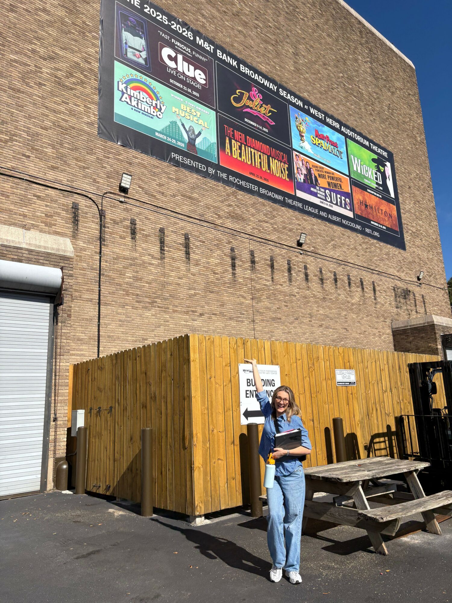 Person standing outside a building with a large signboard, wooden wall, and outdoor seating area, under a clear blue sky.