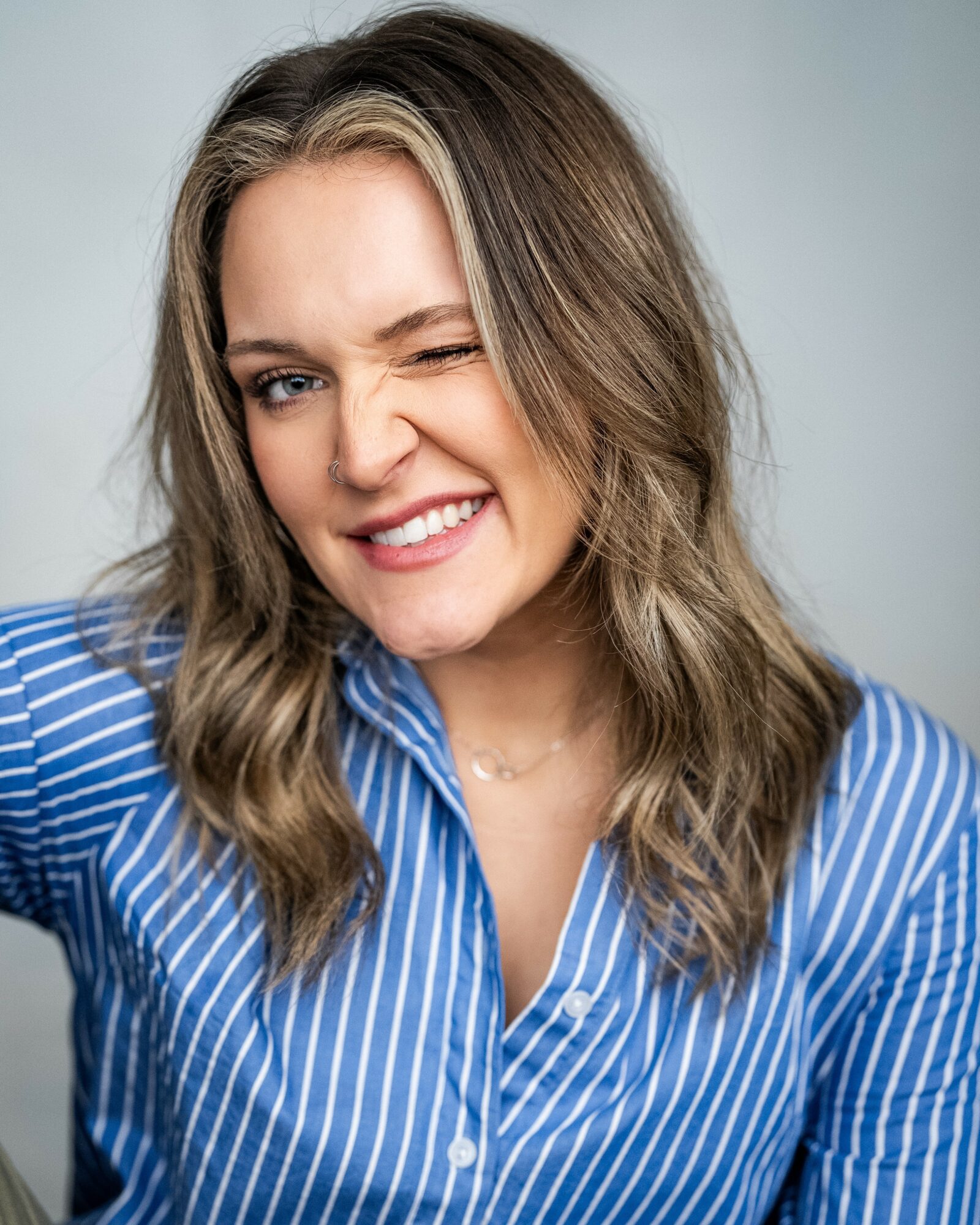 Woman with wavy hair winking and smiling, wearing a blue and white striped shirt, against a plain background.
