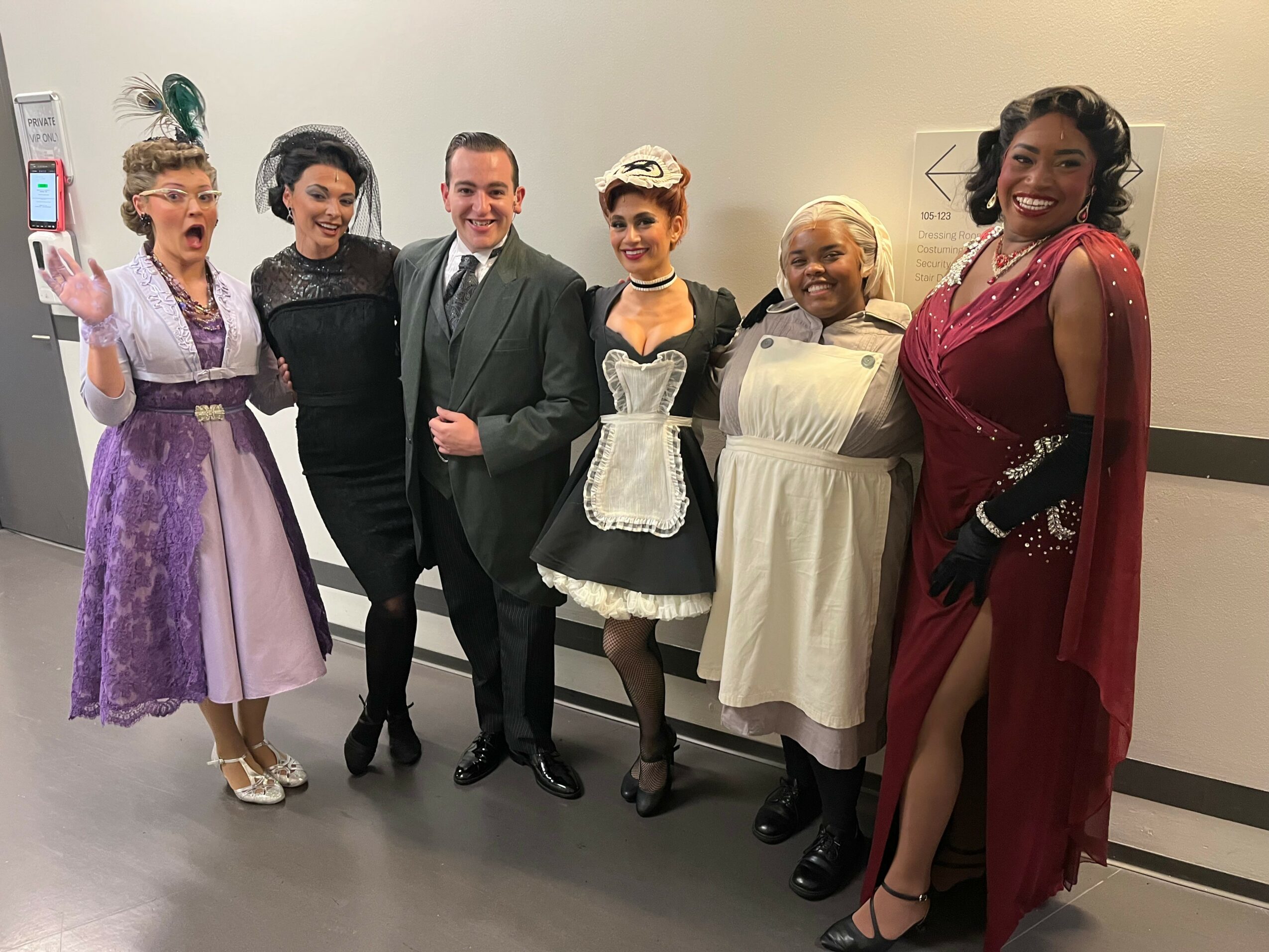 Six people in costumes and formal attire pose together indoors, smiling for the camera.