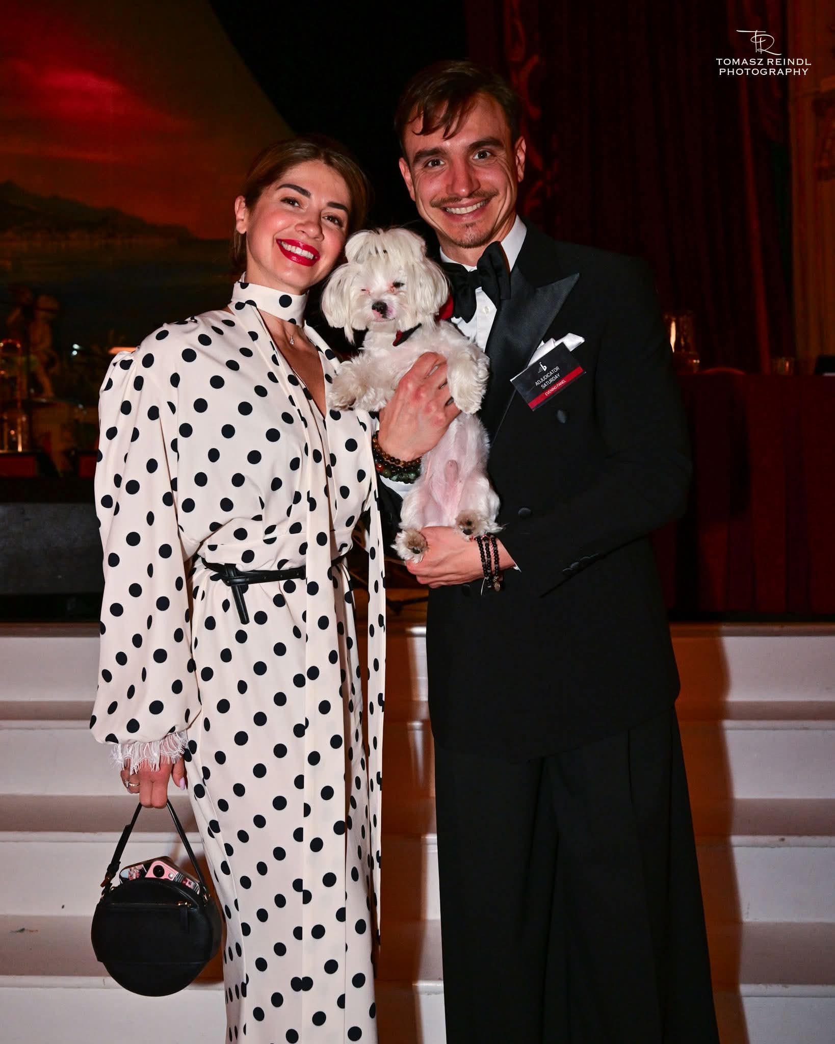 Two people smiling, holding a small white dog, indoors with a dark background and red curtains.