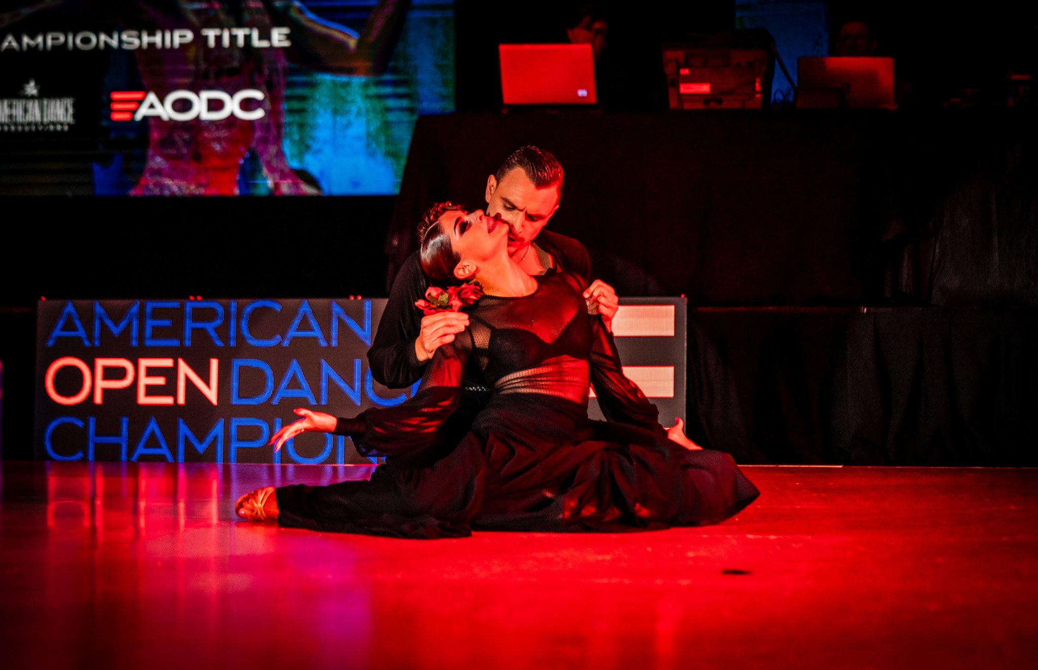 A dancer in a black and red costume performs on stage with a sign reading 'American Open Dance Champion' behind her.