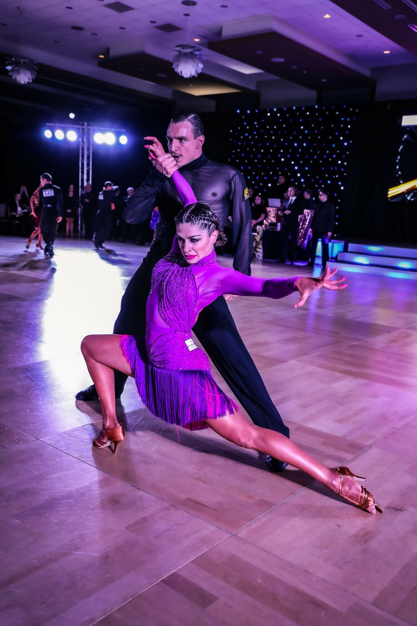 A couple dancing on a wooden floor in a ballroom with purple lighting, the woman in a purple dress and the man in black.
