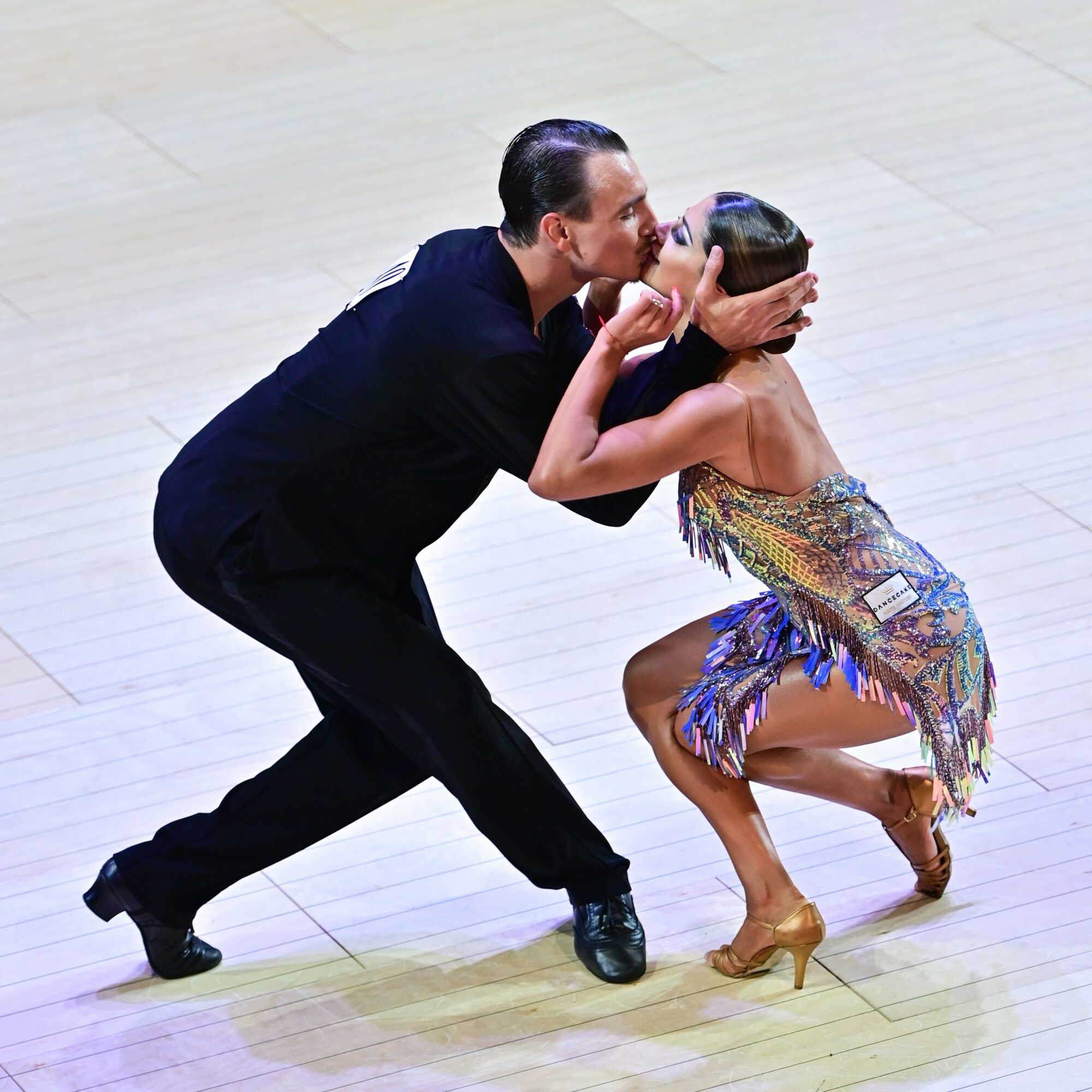 A man and woman dance closely, with the man leaning in and the woman crouching, both in colorful dance attire.