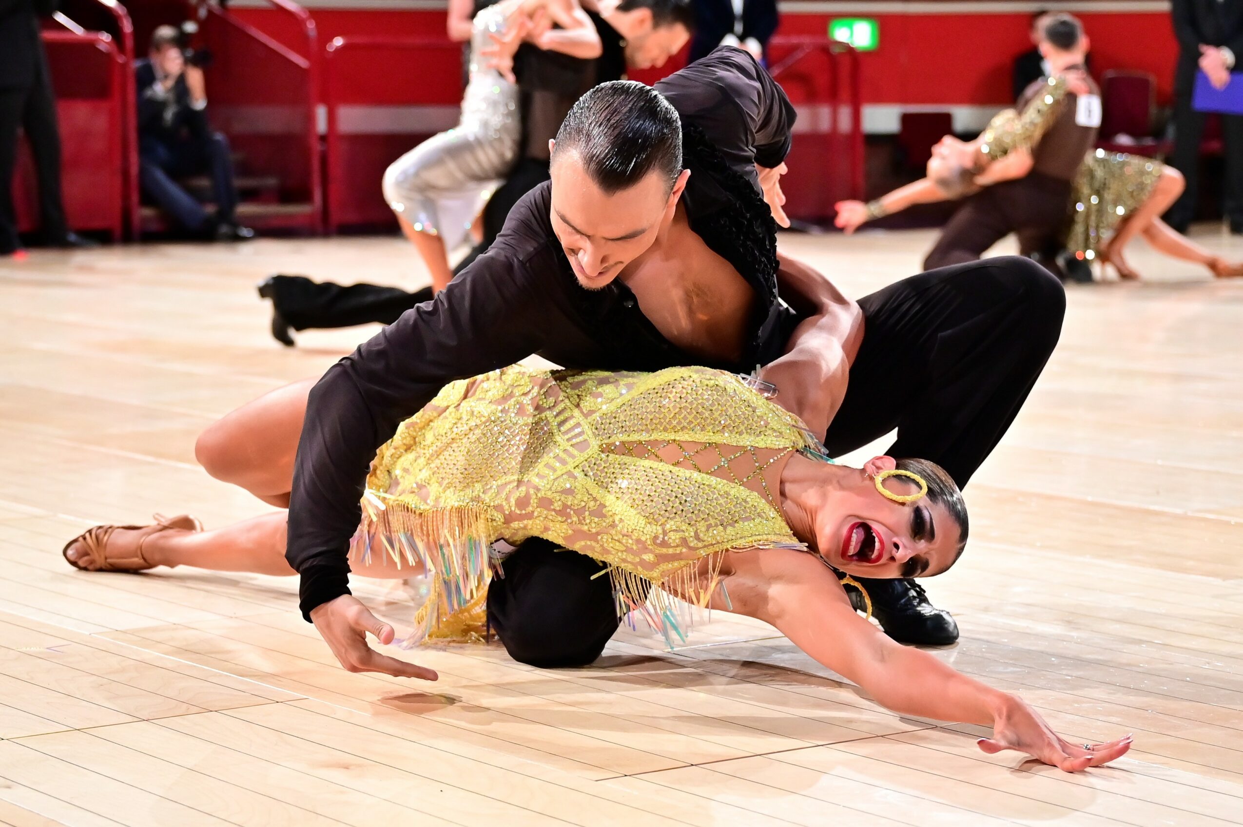 Two dancers perform on a wooden floor, one in a black outfit and the other in a yellow dress, in a dance studio.