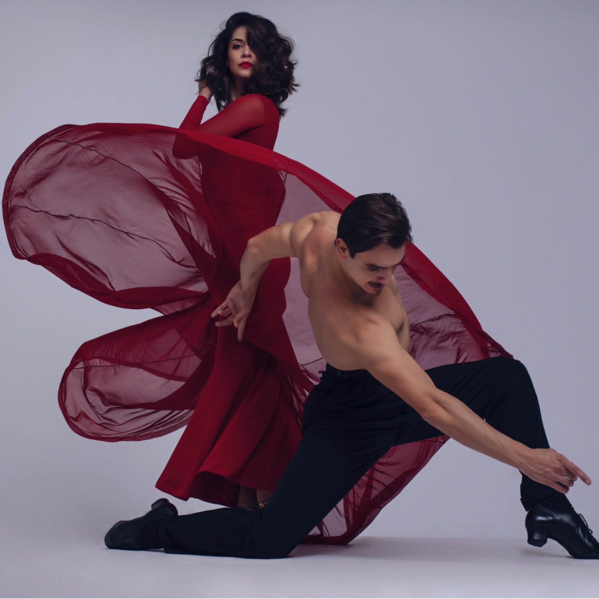 A woman in a red dress and a man in black pants pose dynamically against a plain background.
