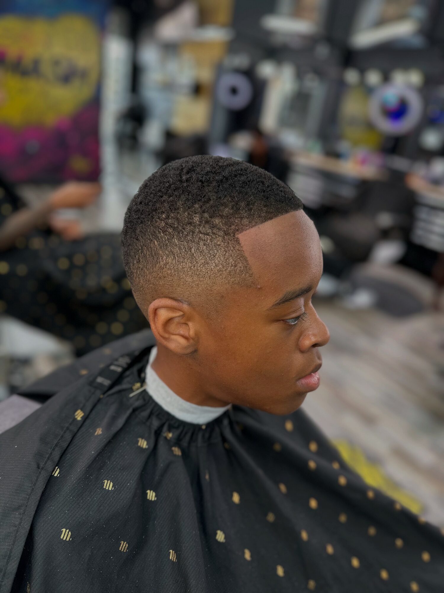 Young man with a short fade haircut, sitting in a barbershop, wearing a black cape with gold dots, looking to the side.