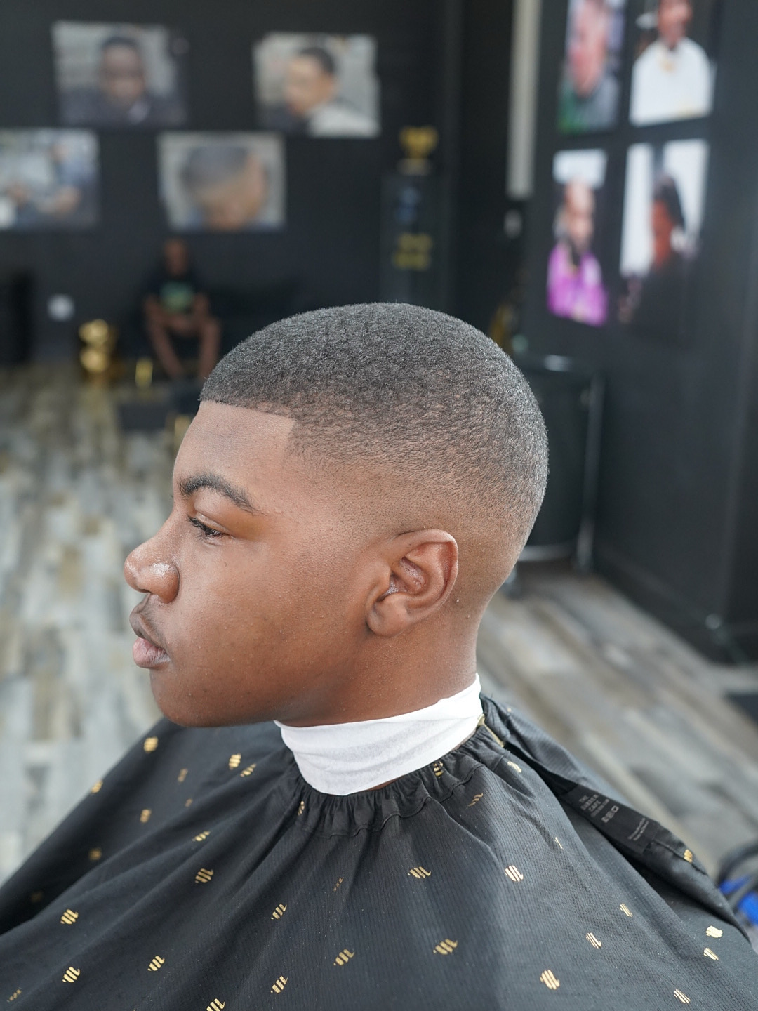 Young man with a short, faded haircut sitting in a barbershop, wearing a black cape with gold accents, background with photos and barber tools.