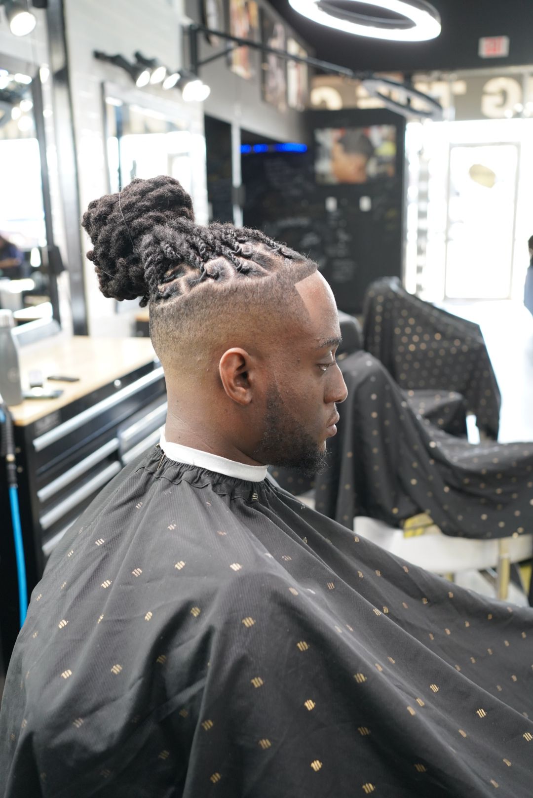 Person with styled hair sitting in a salon chair, wearing a black cape with gold dots, in a modern salon setting.