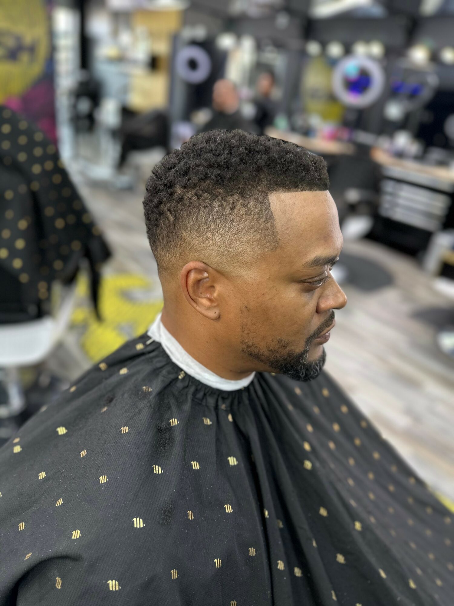 Side profile of a man with a fresh fade haircut in a barbershop, wearing a black cape with gold dots.