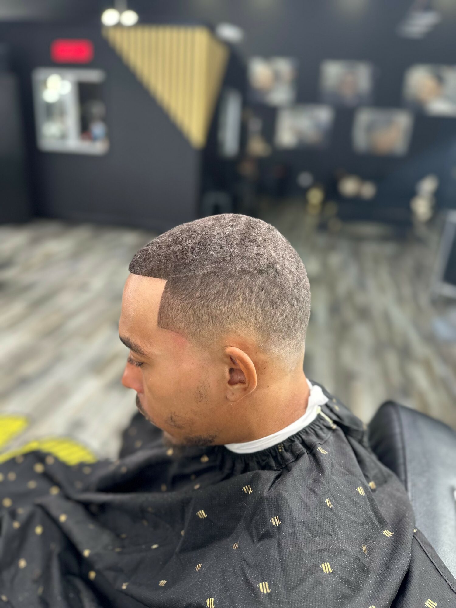 Man with a short, faded haircut sitting in a barbershop chair, black cape with small yellow squares, blurred background