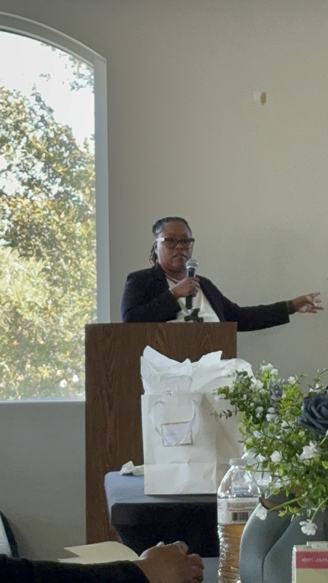 Person speaking at podium with window and trees in background, table with flowers and gift bags in foreground.