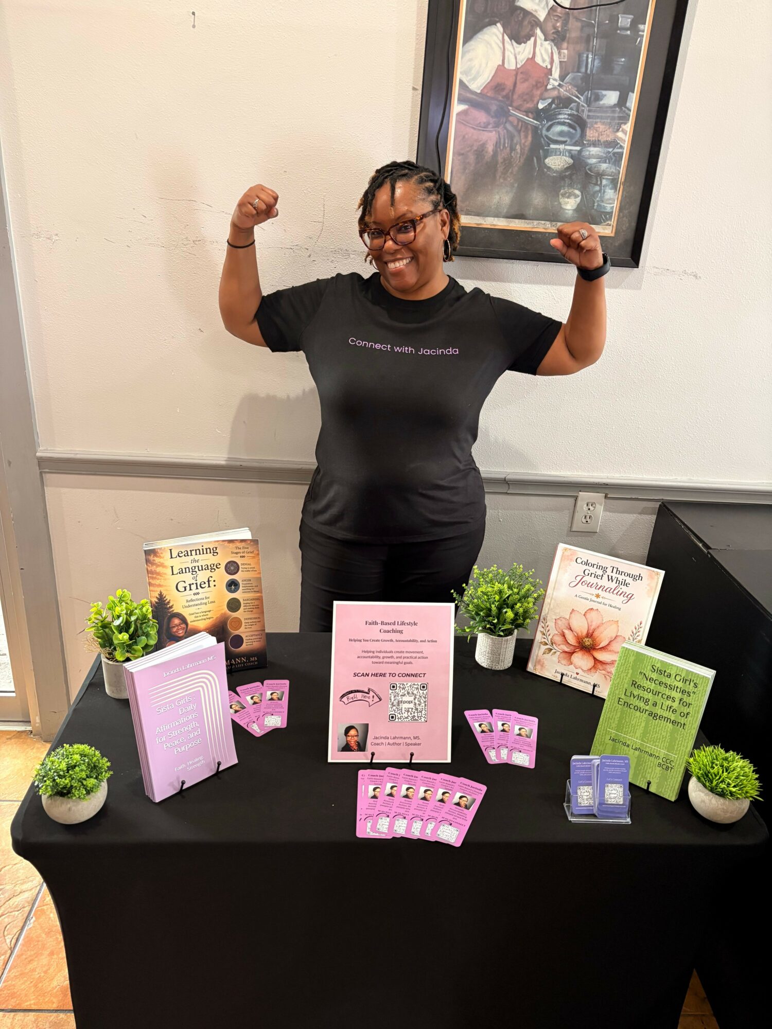 Person standing next to a table with pamphlets, plants, and a pink box, smiling and flexing arms.
