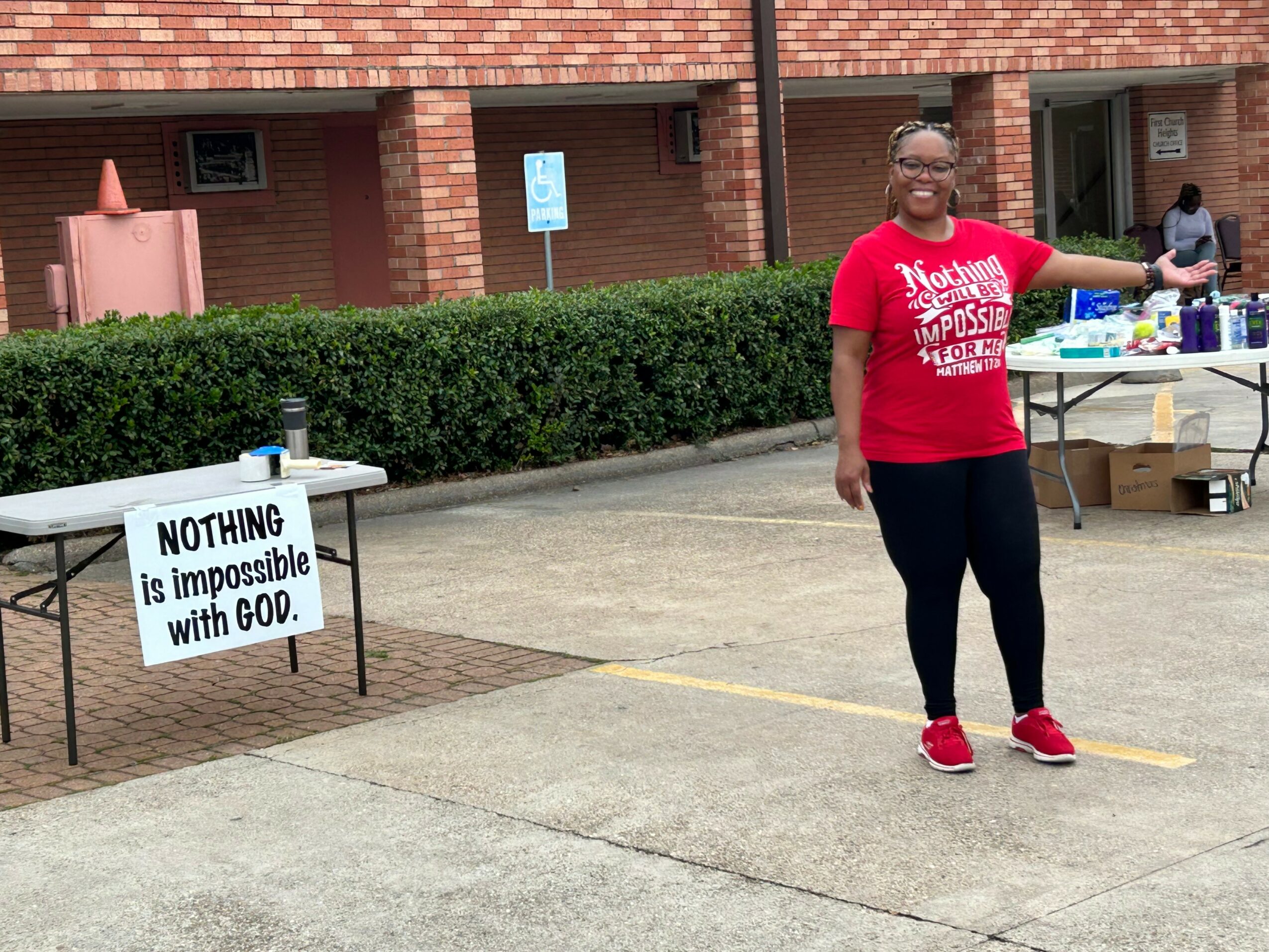 Woman standing outdoors near a table with a sign that reads 'Nothing is impossible with God,' smiling and pointing to the right.